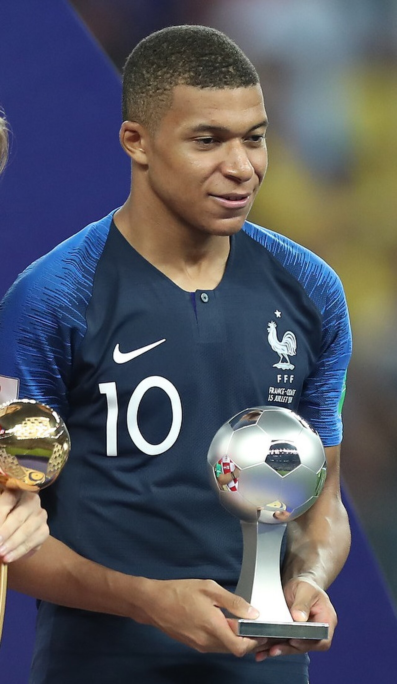 Kylian Mbappé in a blue soccer jersey holding a trophy