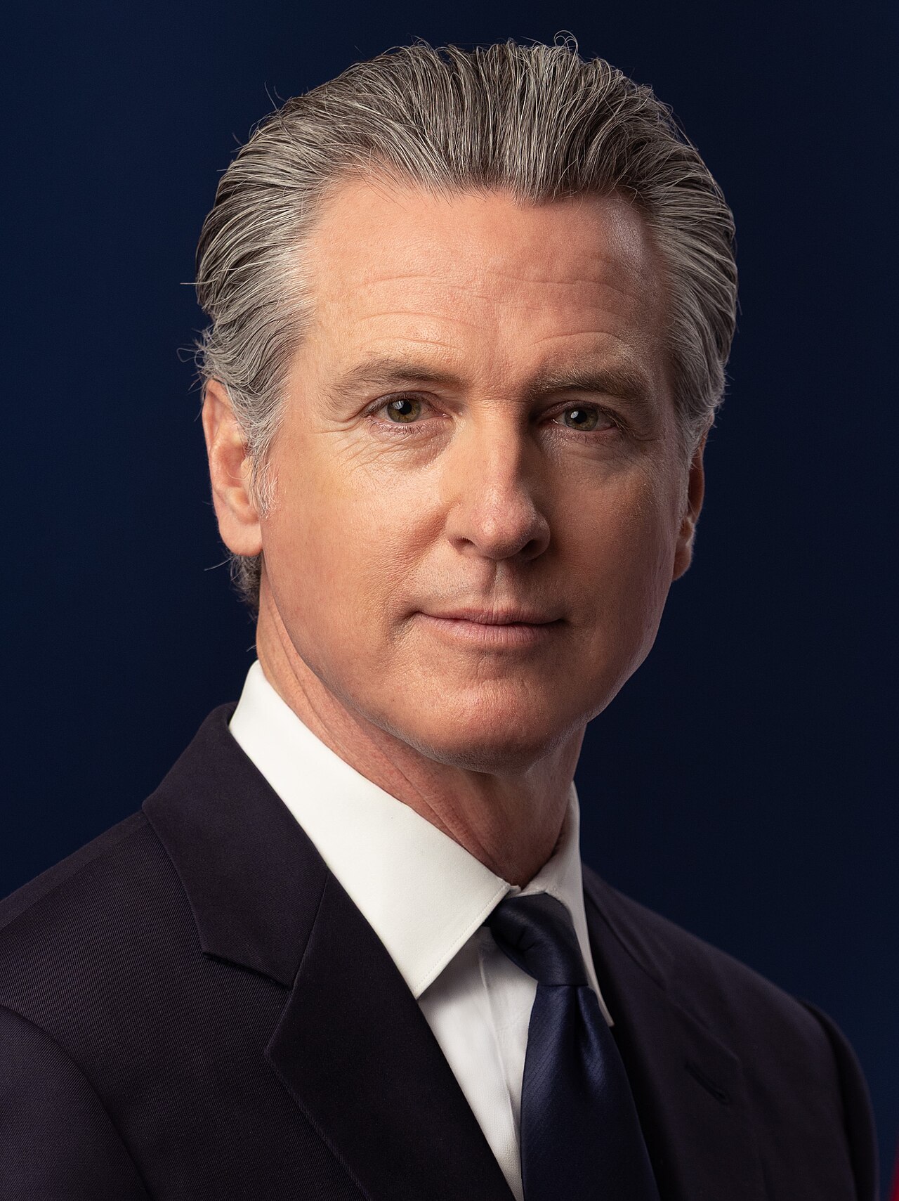 Gavin Newsom, a middle-aged man with graying hair, wearing a dark suit and tie, against a dark blue background.