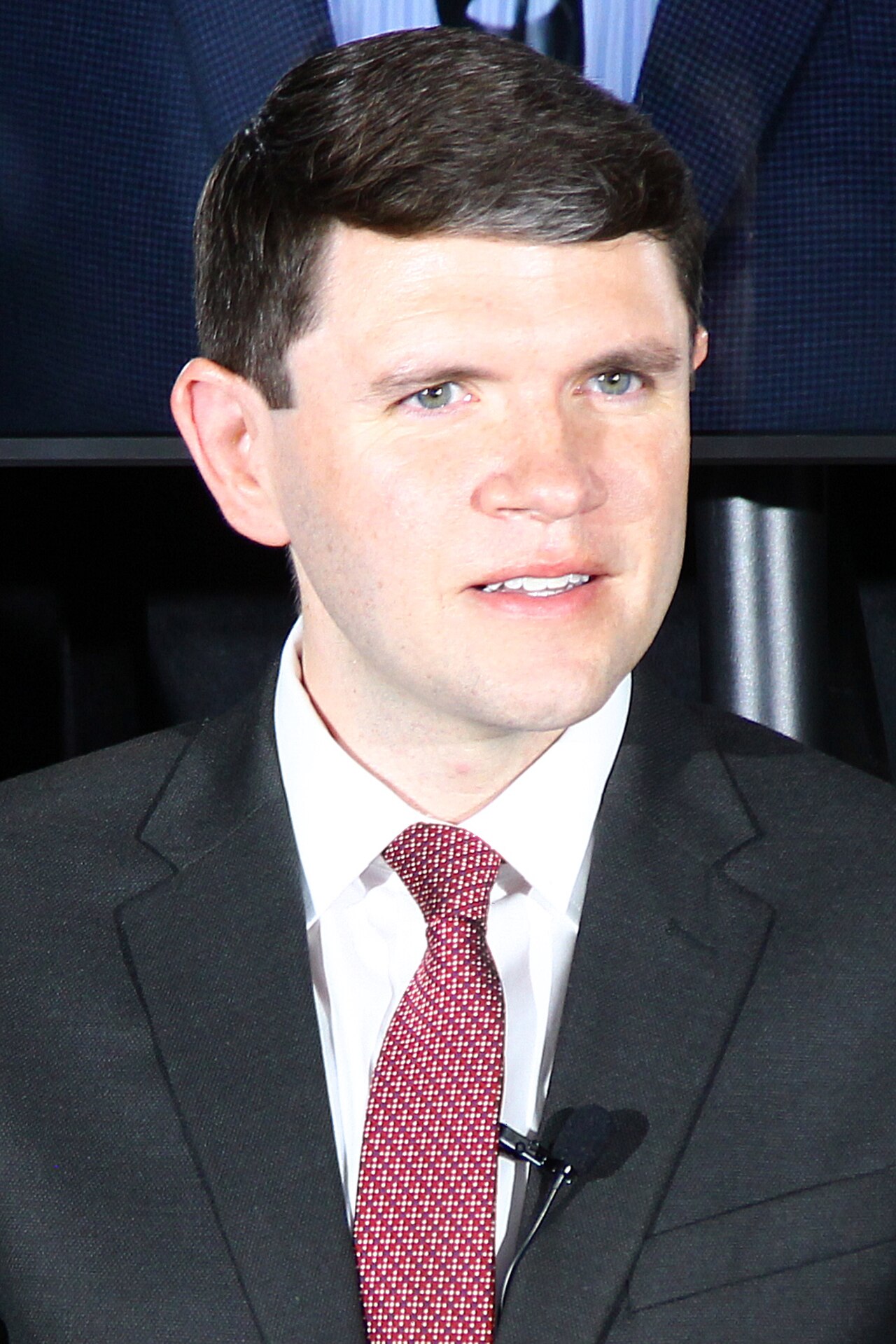 A man with short brown hair, wearing a black suit, white shirt, and red tie with white dots, and a small microphone clipped to his tie.