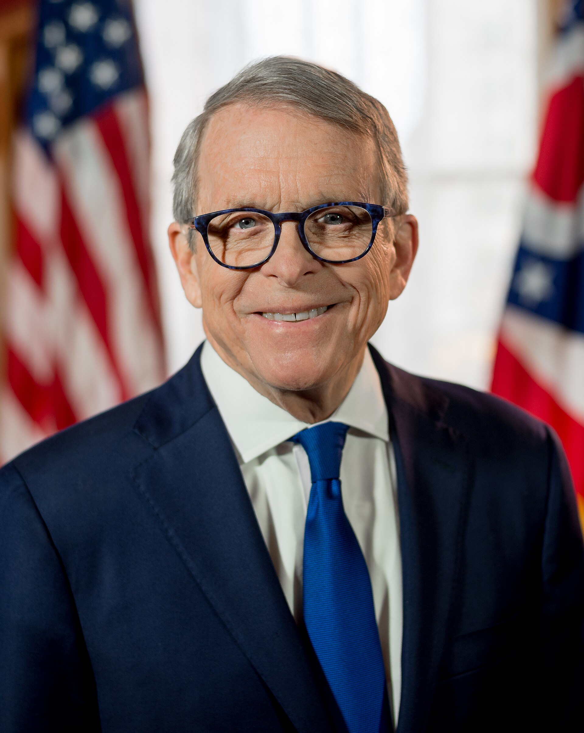 A portrait of Ohio Governor Mike DeWine, a white man with graying hair, wearing glasses, a dark suit, white shirt, and blue tie, standing in front of two American flags.