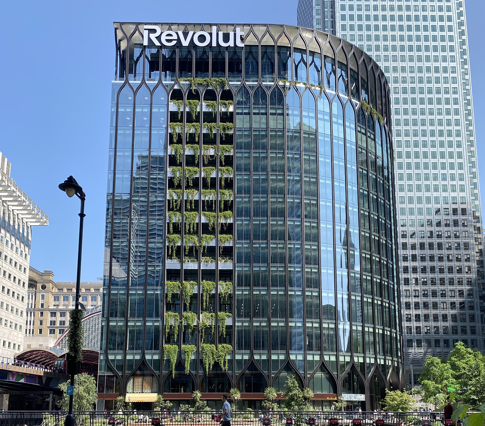 A modern glass-fronted office building with the Revolut logo prominently displayed at the top. The building features vertical green planters cascading down sections of its facade.