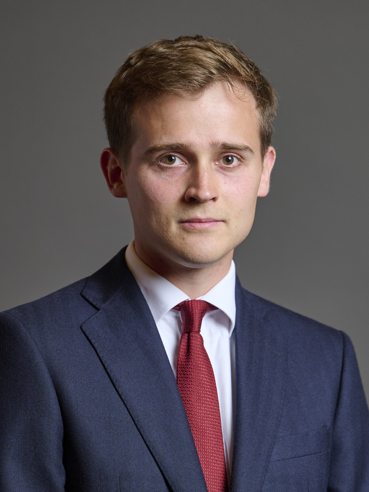 A fair-skinned man with short brown hair wearing a blue suit, white shirt, and red tie.