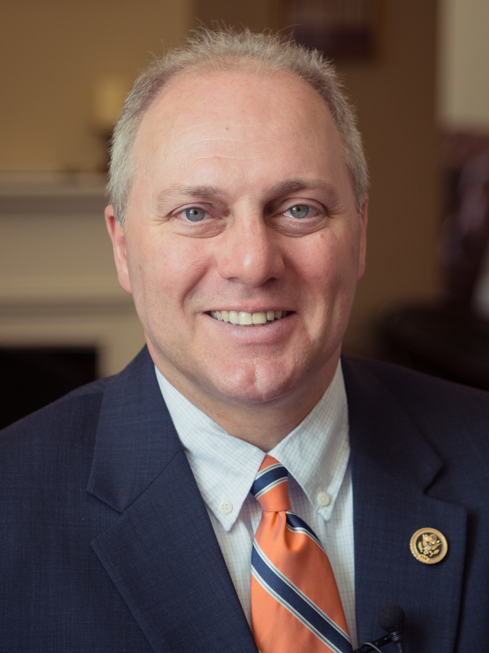 A smiling middle-aged man with short gray hair, wearing a blue suit, white shirt, and orange tie with blue stripes, and a gold pin on his lapel, looking directly at the camera.