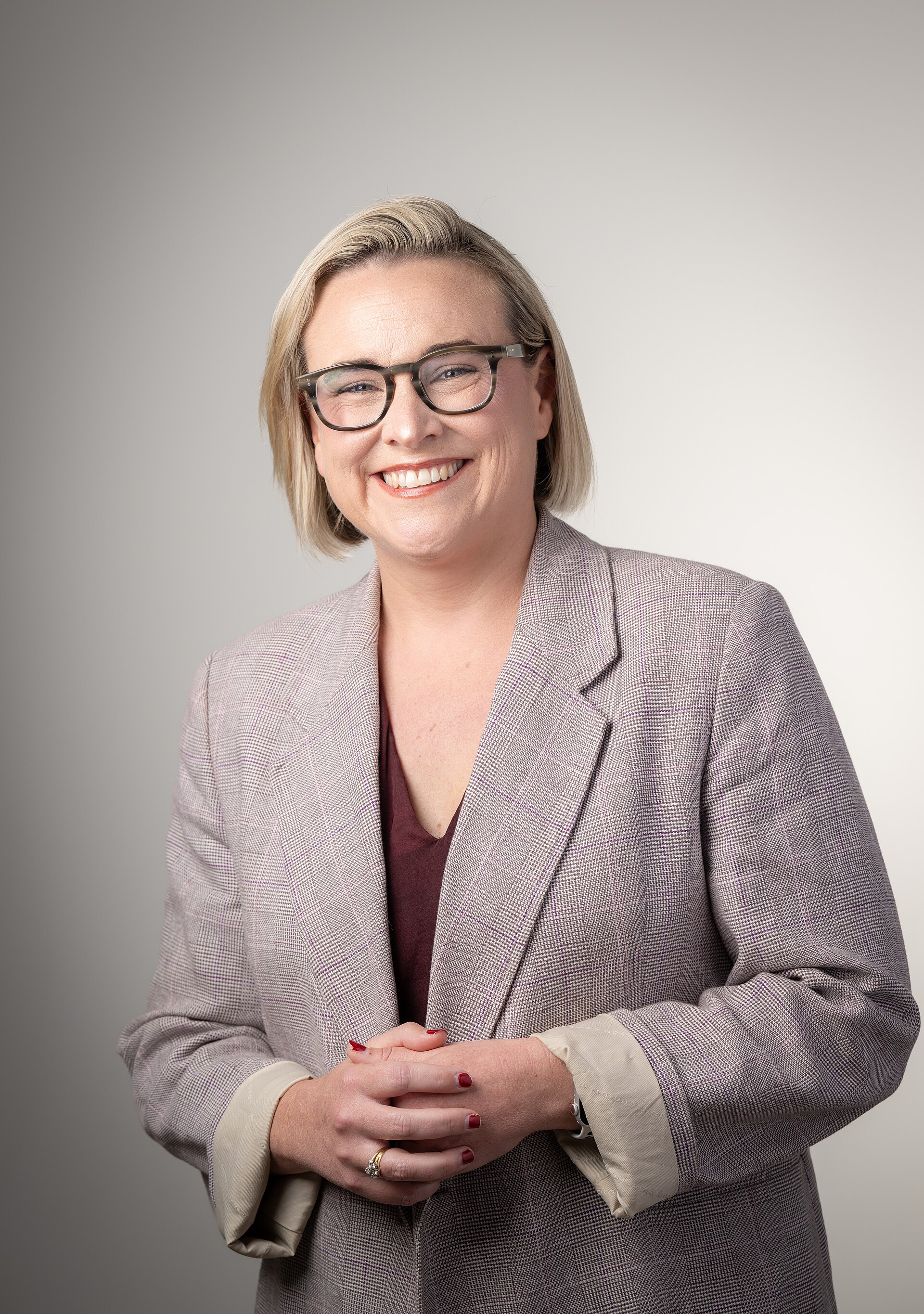A smiling middle-aged woman with short blonde hair, wearing a gray blazer, a maroon top, and black-framed glasses, with her hands clasped in front of her.