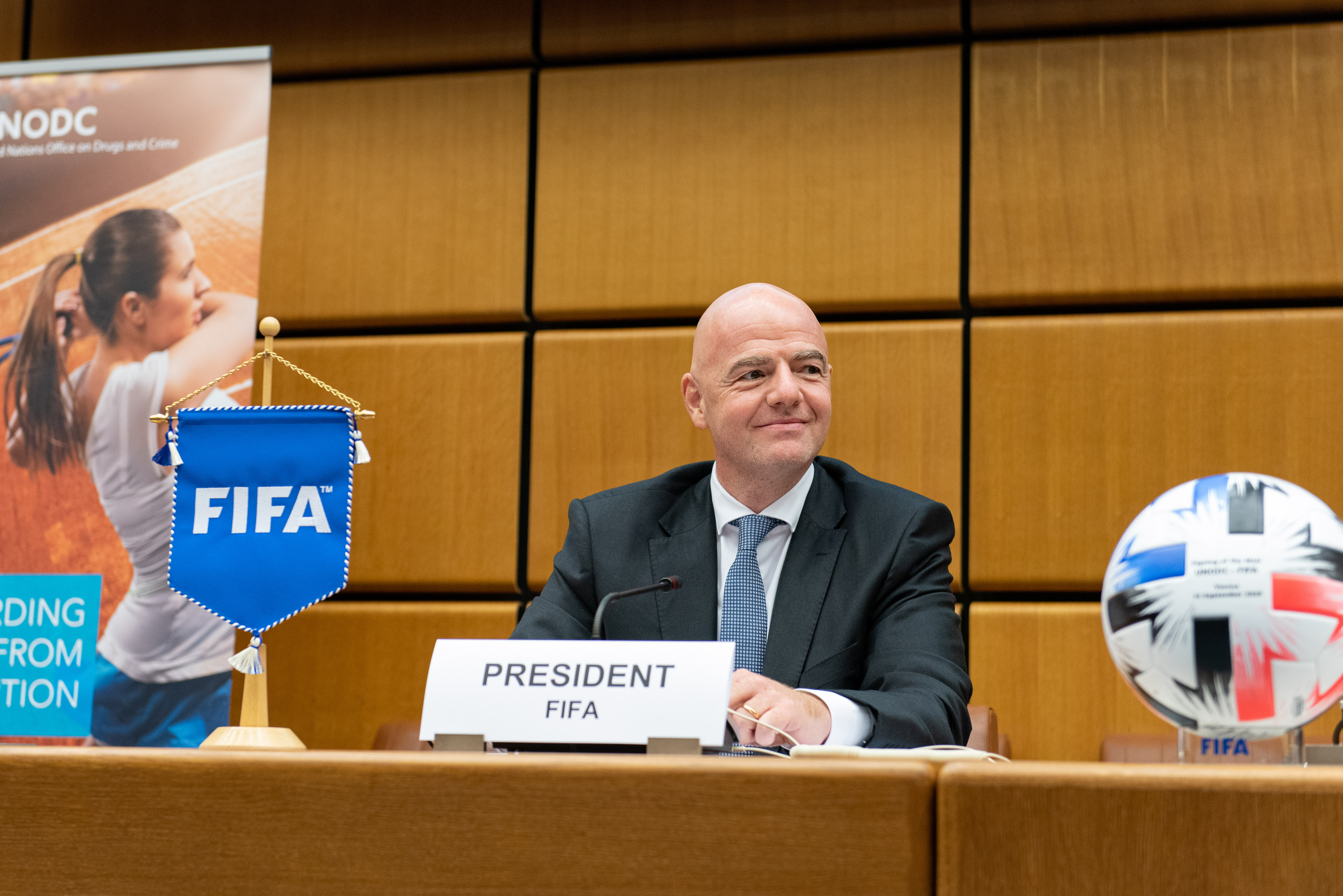 Gianni Infantino sits behind a FIFA president placard, with a FIFA pennant and soccer ball beside him.