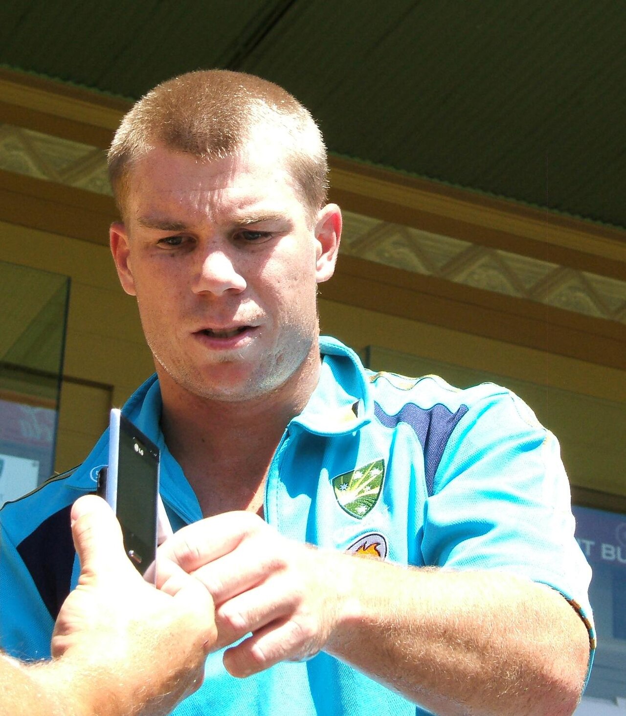 A man with short blond hair wearing a light blue polo shirt with a logo on the left side of his chest, holding a cell phone in his right hand and looking down at it with his left hand in front of him.