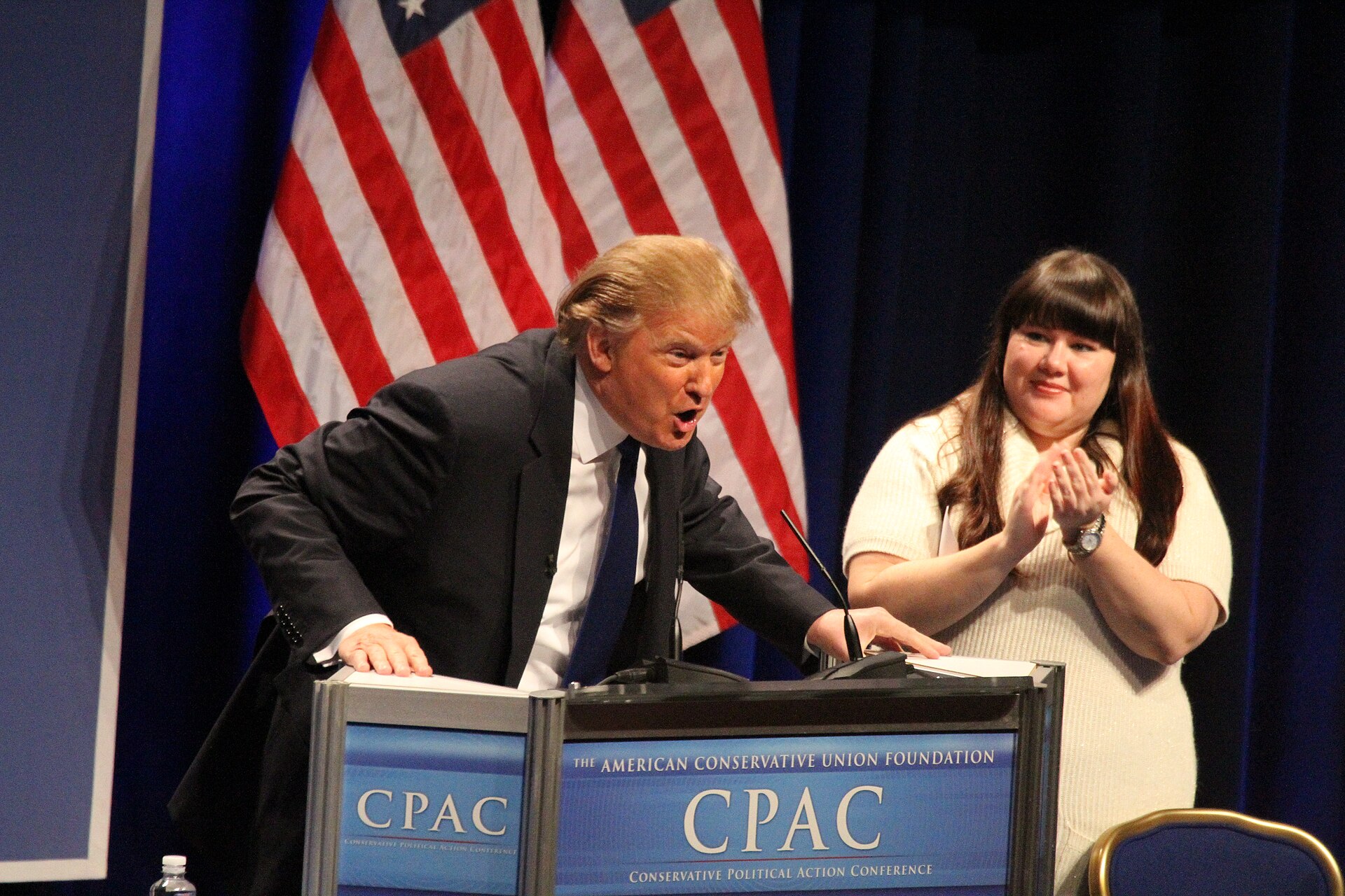 Donald Trump speaks at a podium with "CPAC" and "The American Conservative Union Foundation" visible, while a woman applauds behind him.