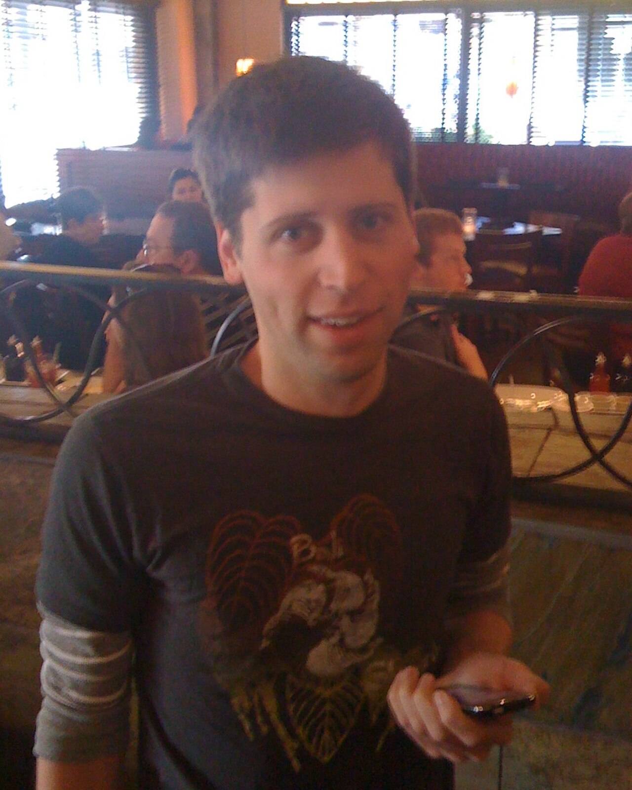 A man with short brown hair wearing a dark gray t-shirt with a graphic on it, holding a cell phone in his right hand and smiling at the camera.