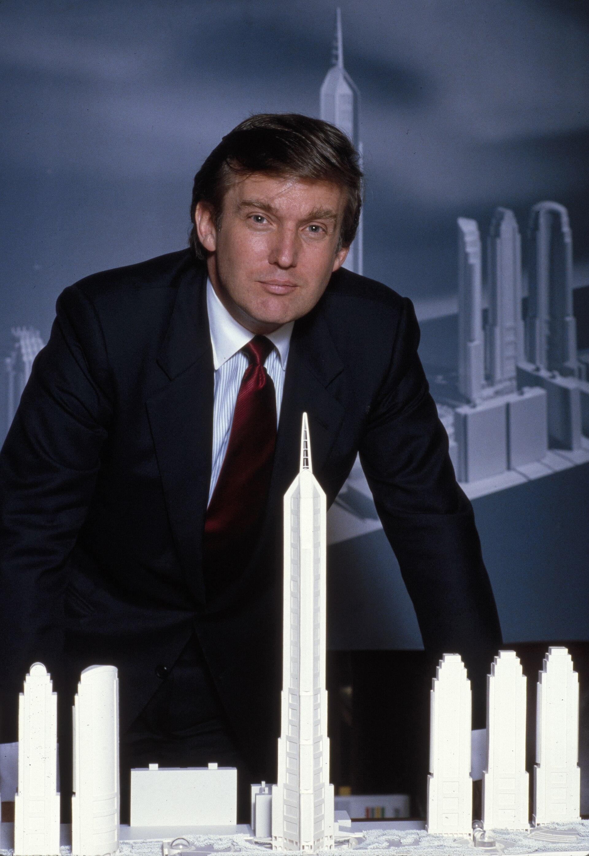 Donald Trump in a black suit and red tie leaning on a table with a model of skyscrapers