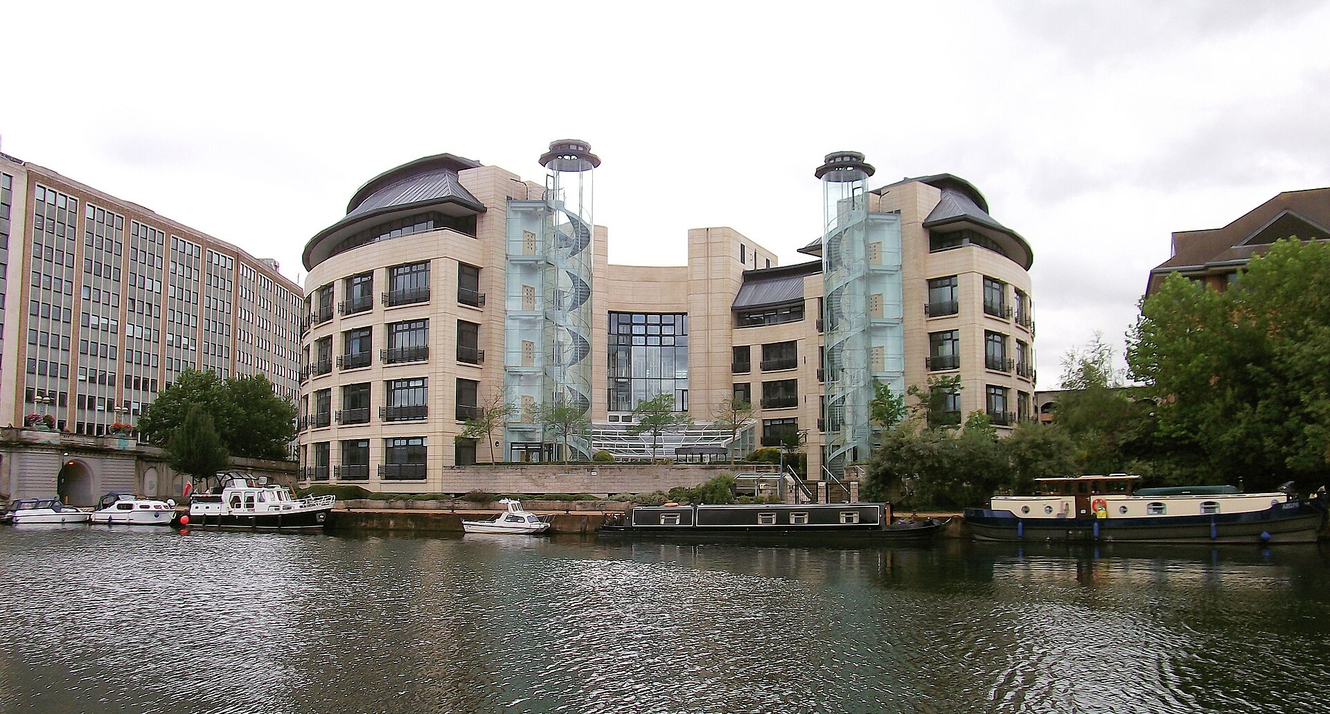 A wide shot of a river with several boats docked along the bank, and modern buildings on the opposite side.