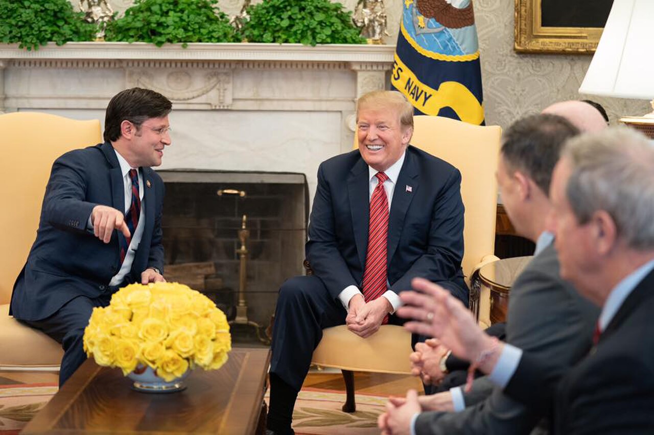 Archival photograph from March 5, 2019: President Donald Trump seated in the Oval Office with Mike Johnson; a U.S. Navy flag and yellow roses are visible.