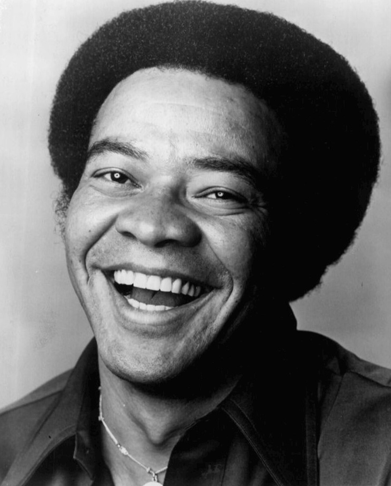 Bill Withers smiling and looking at the camera, wearing a collared shirt and a necklace with a pendant, against a plain background.