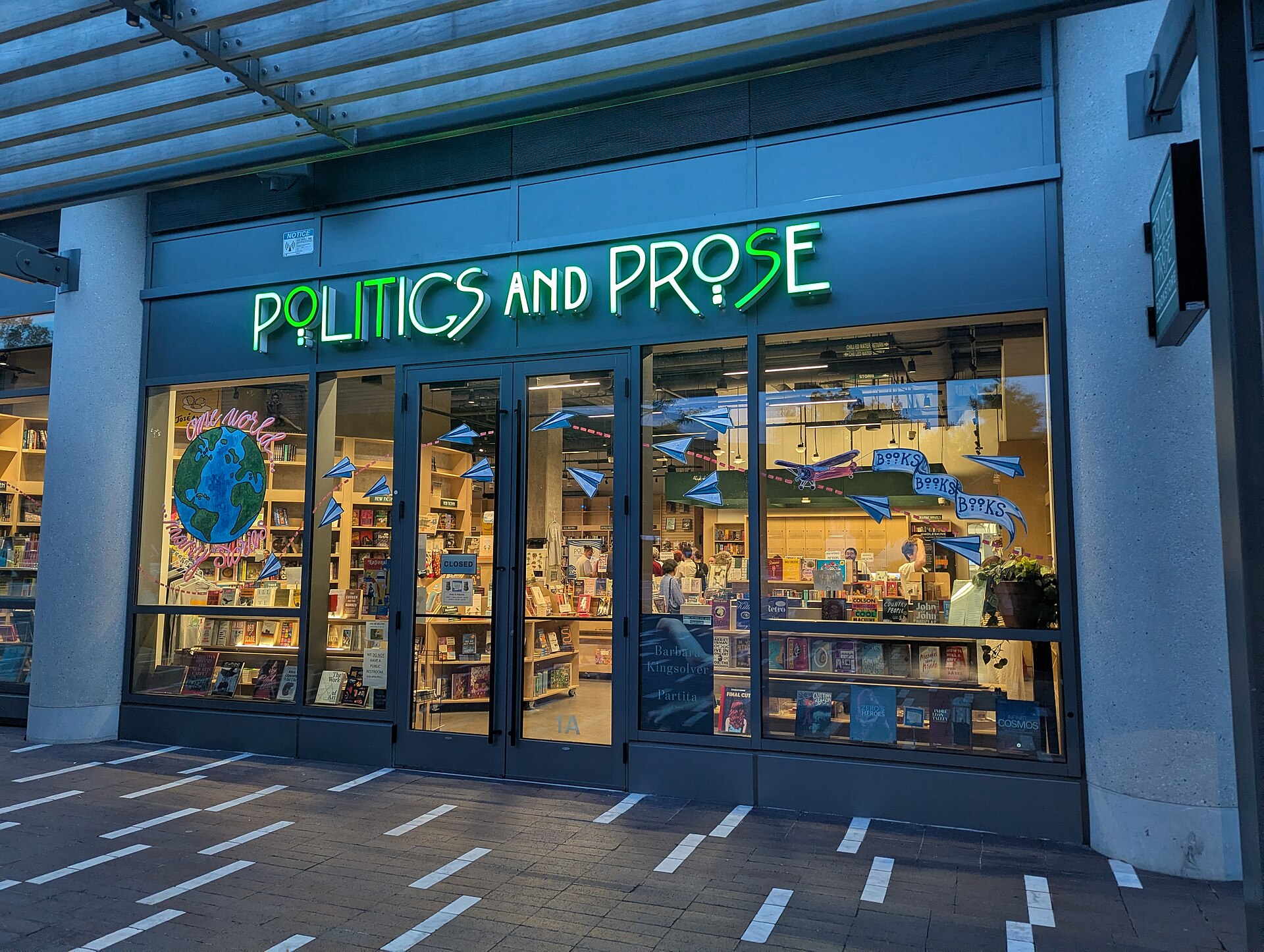 The storefront of Politics and Prose bookstore, with its name illuminated in neon green letters above the entrance. The windows display books and decorations, including paper airplanes and a globe.