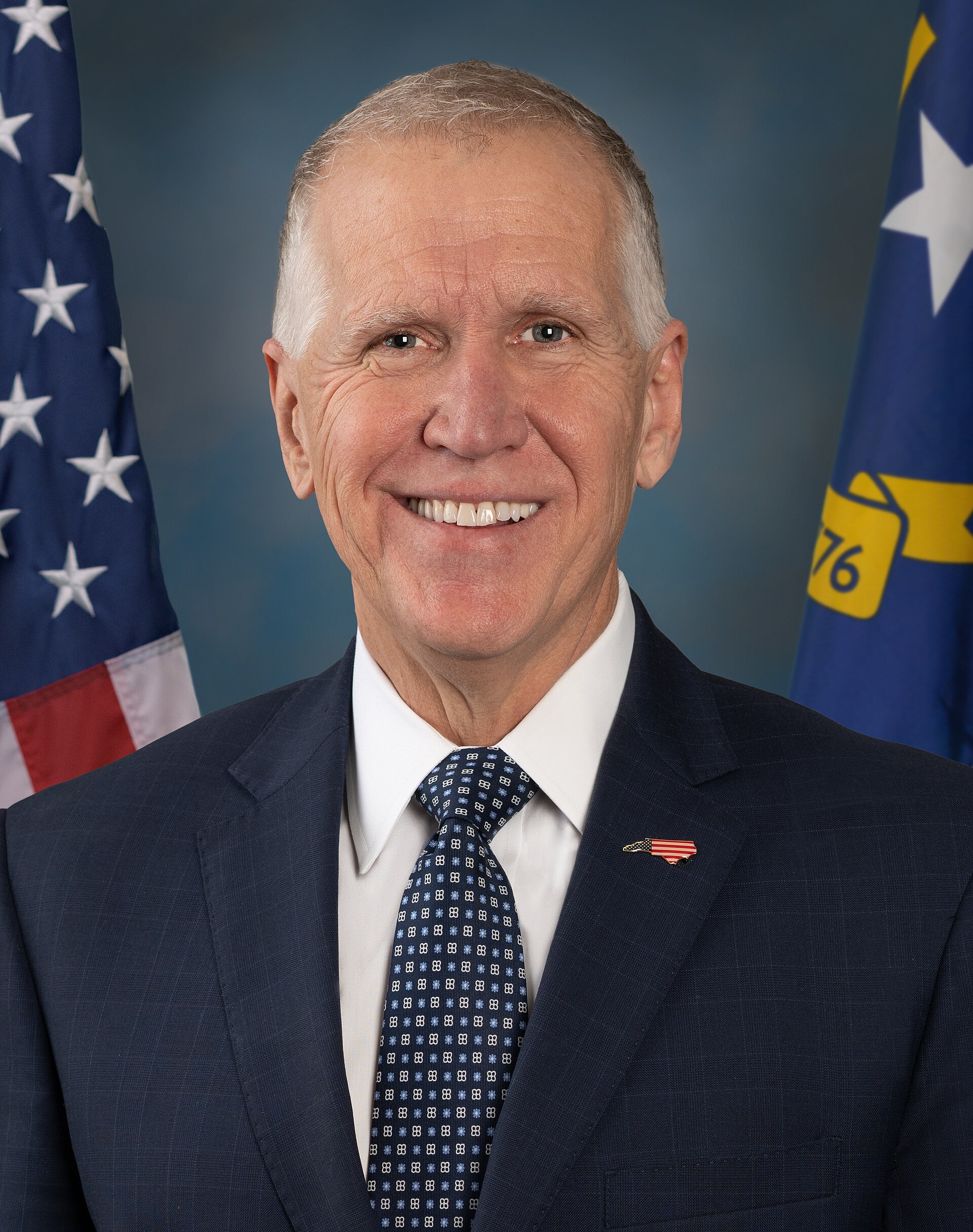 A formal portrait of a smiling white man in a dark suit and blue tie, standing between the American flag and a blue flag with a yellow emblem and the number 76.