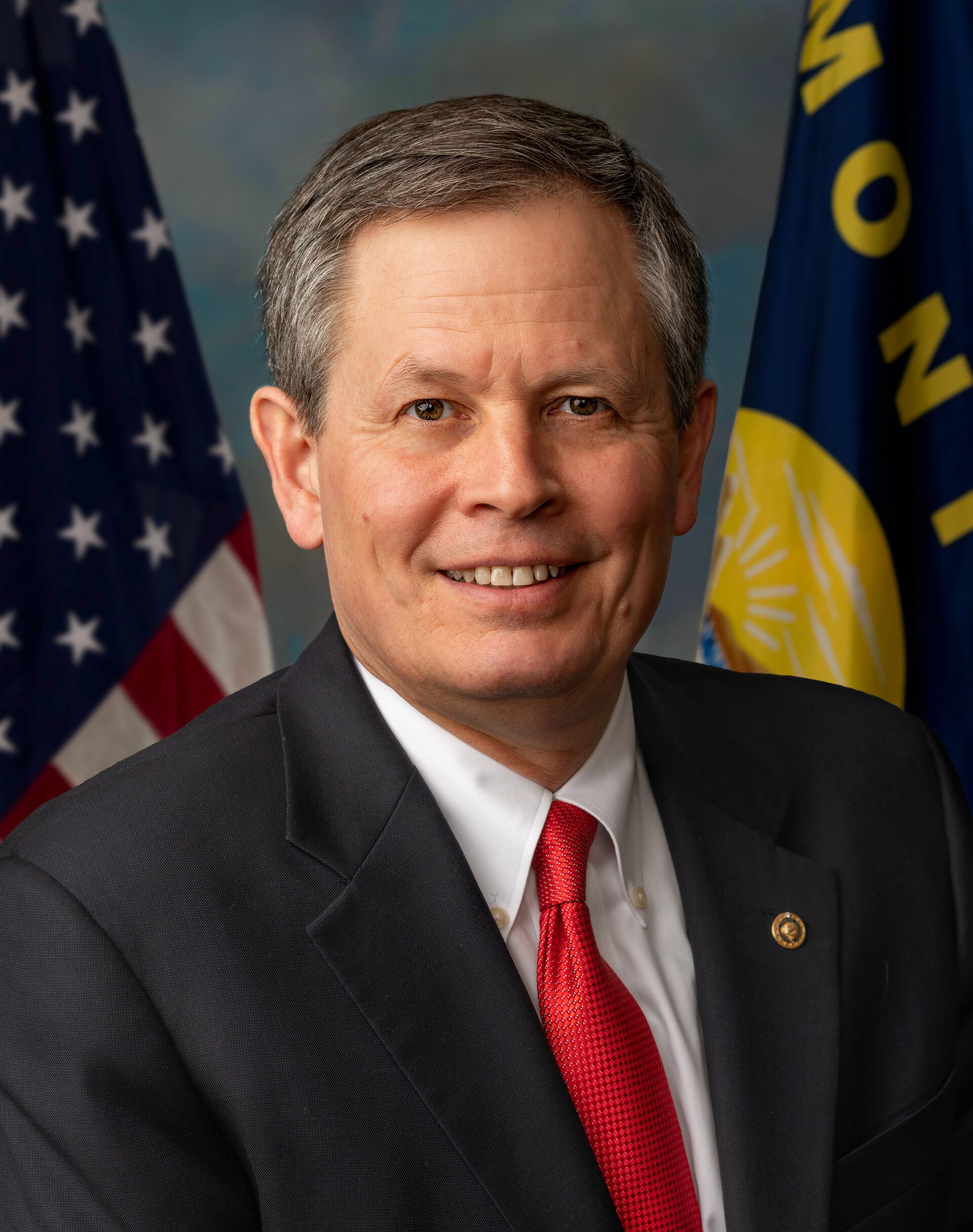 A smiling middle-aged man in a black suit, white shirt, and red tie, with two flags behind him.