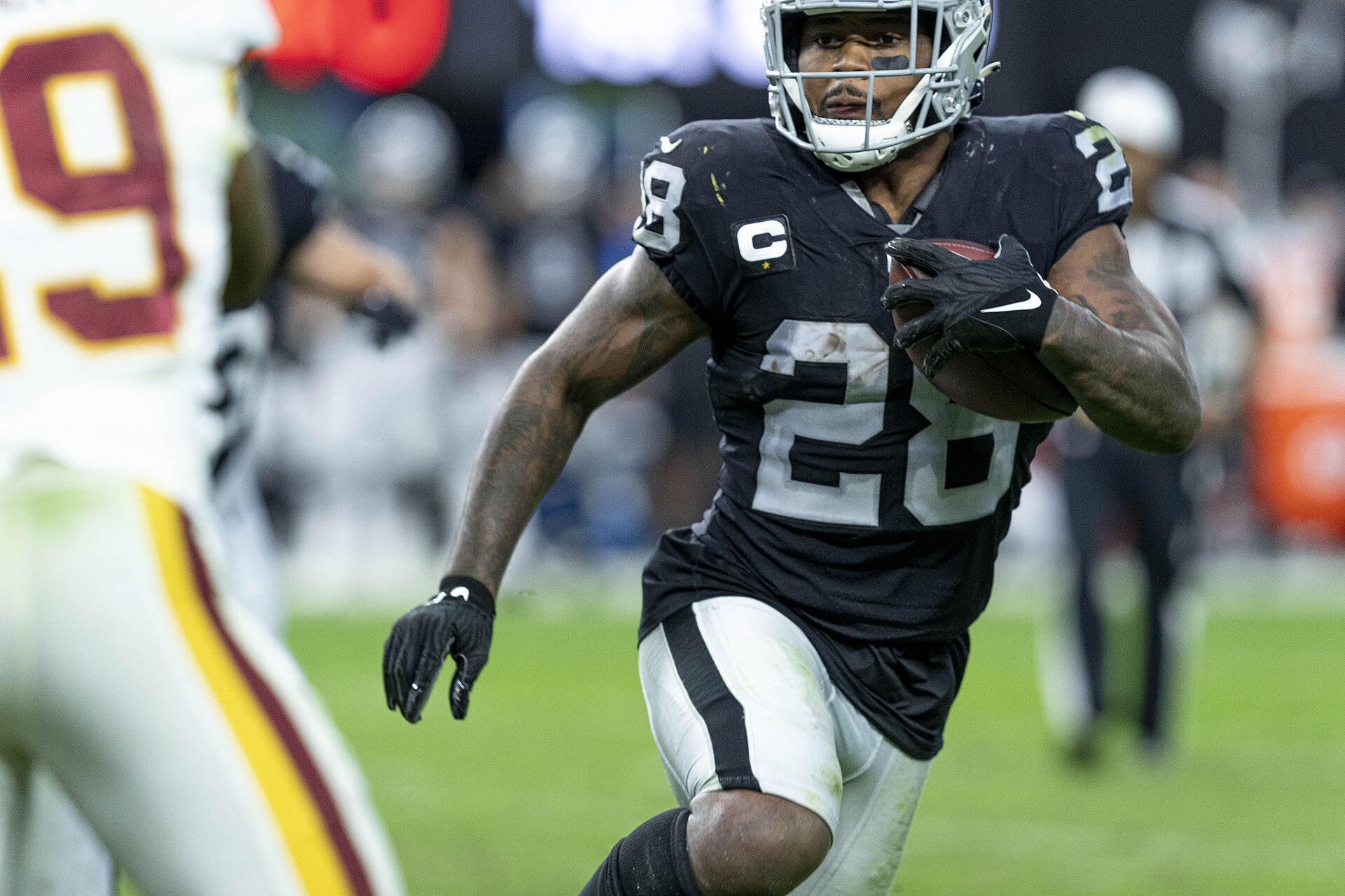 A football player in a black Raiders uniform with the number 28 on his jersey runs with the ball, wearing a helmet and black gloves.