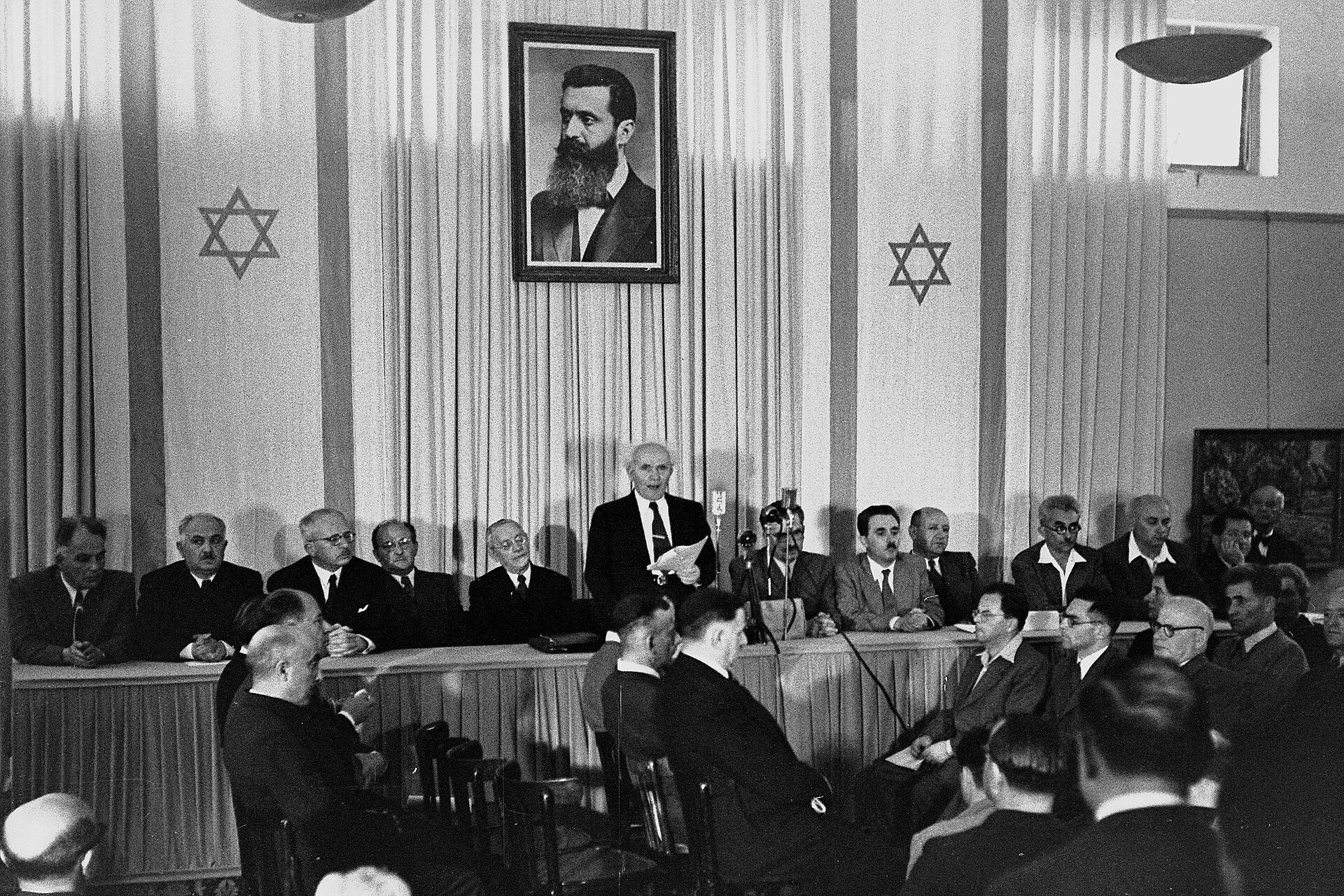A black and white photograph of a formal gathering with men in suits seated and standing. A portrait of a man with a beard hangs on the wall behind a speaker at a podium. Two Star of David symbols are visible on the wall.
