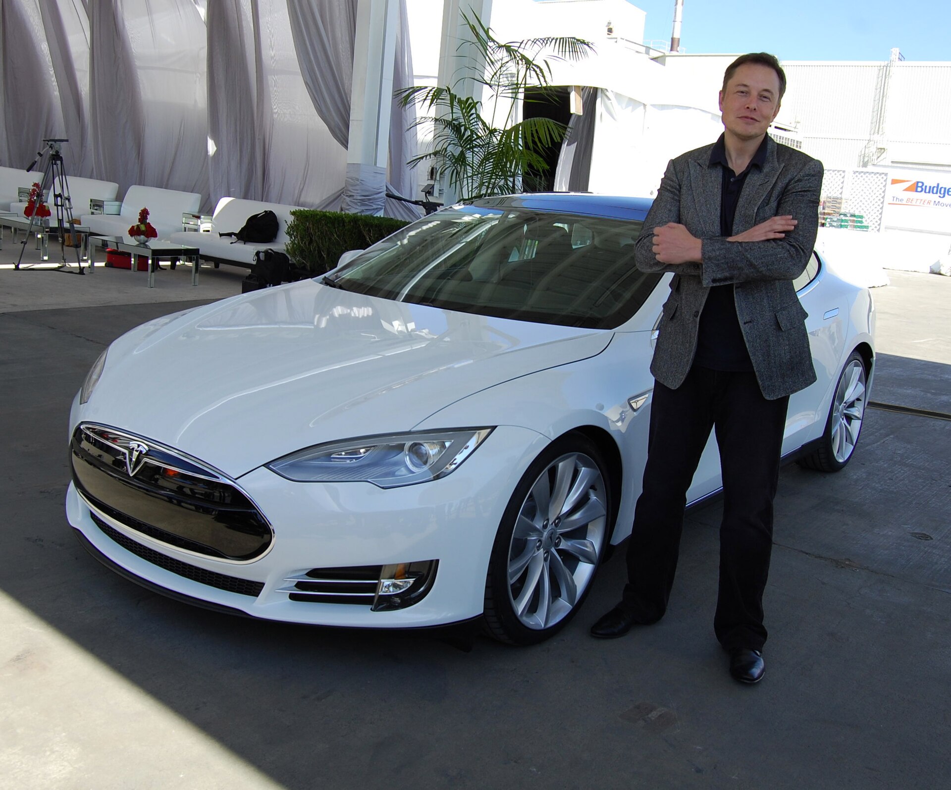 Elon Musk standing with his arms crossed next to a white Tesla car.
