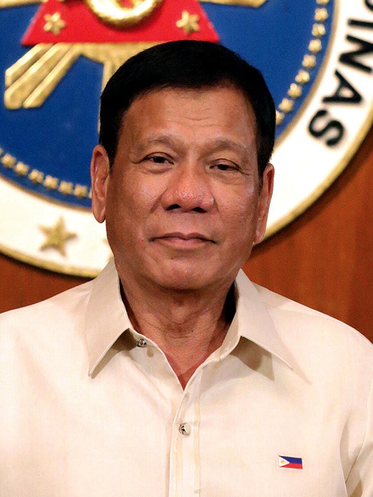 A man with short black hair wearing a white collared shirt with a small Philippine flag pin on the left side of his chest, standing in front of a circular emblem on a wooden wall.