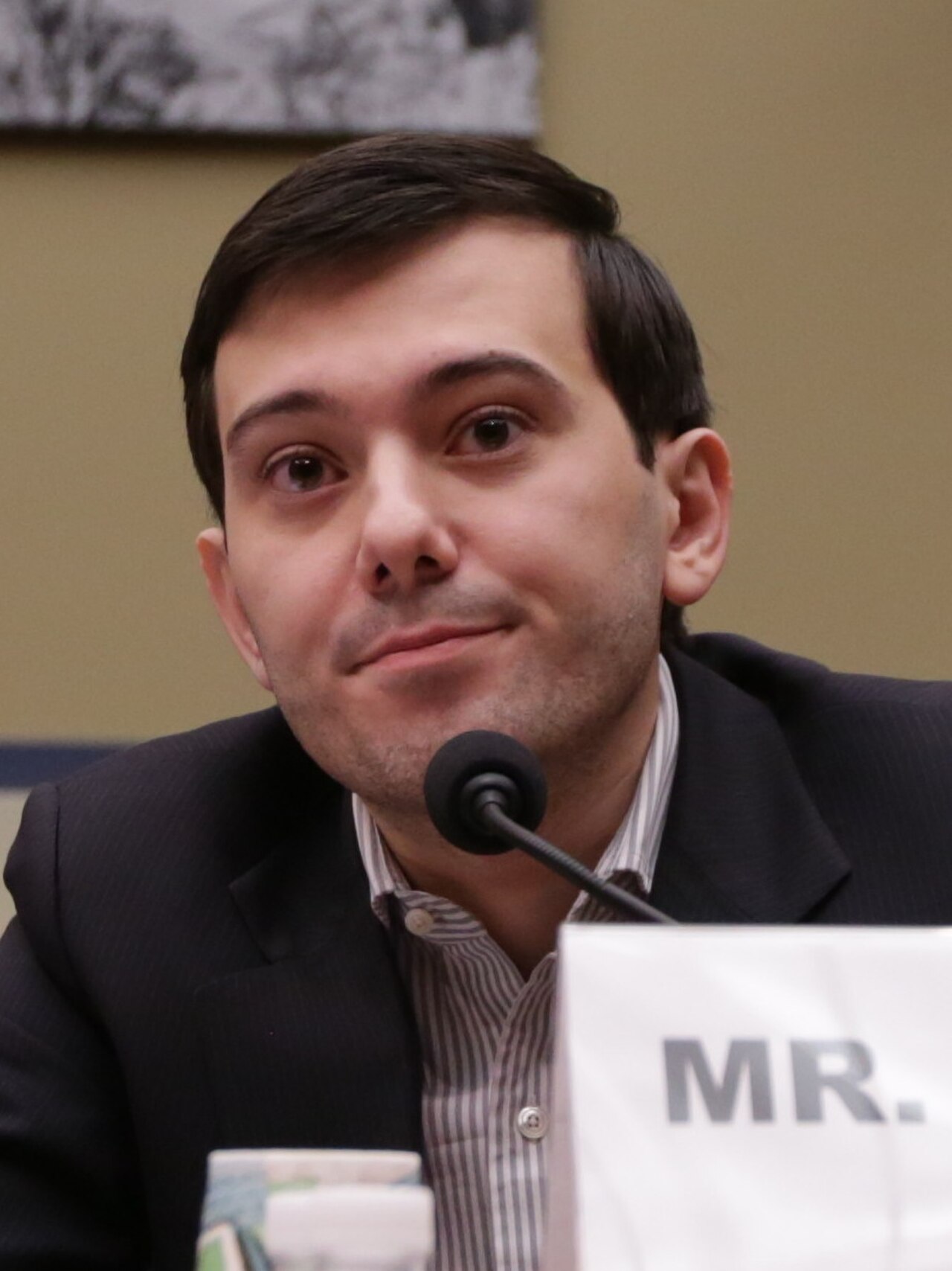 A man with short dark hair wearing a dark suit jacket and striped collared shirt, sitting at a table with a microphone in front of him and a name card that reads 'MR.'