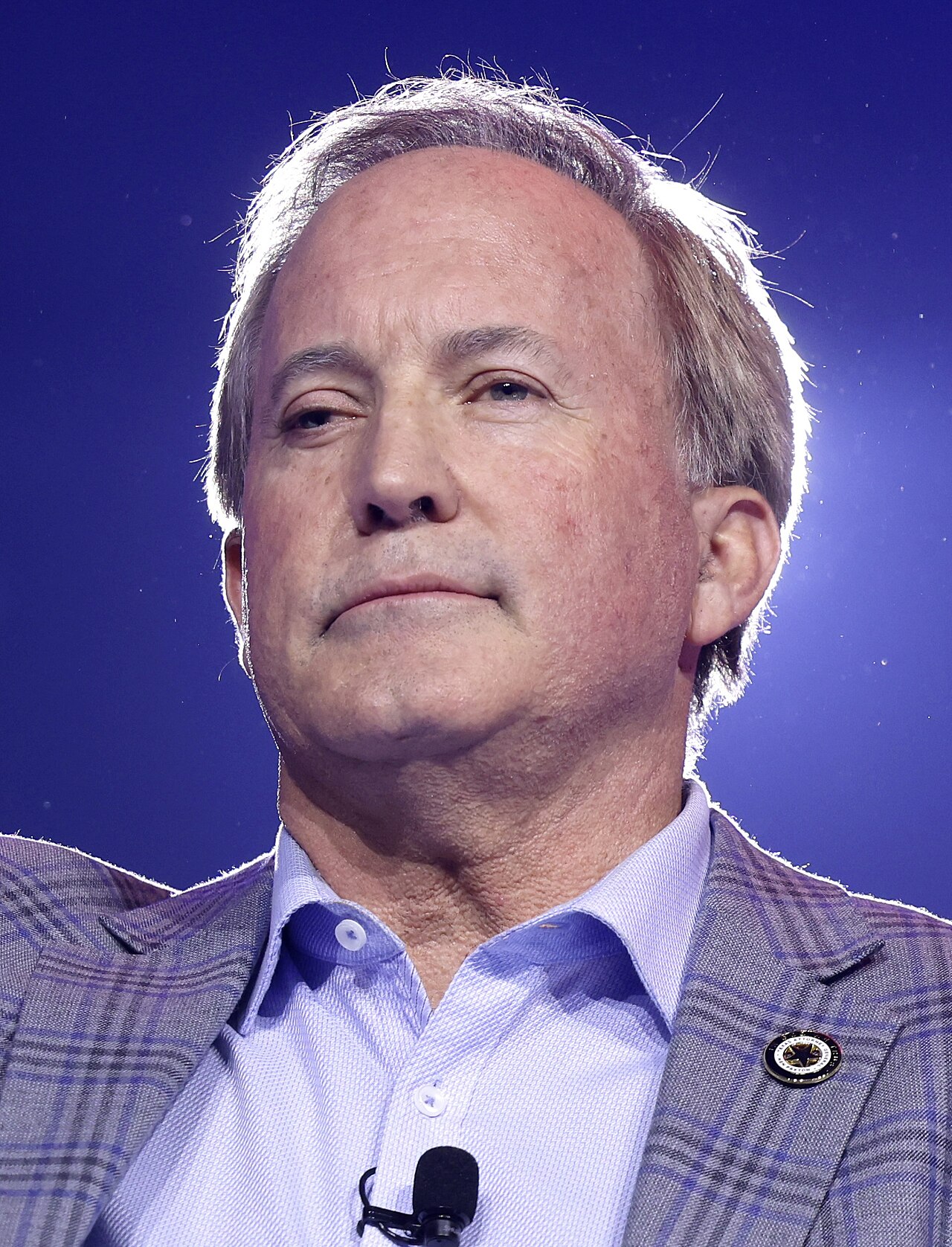 A man with short gray hair wearing a blue shirt and gray plaid jacket with a pin on the lapel, looking to the left with a small microphone visible at the bottom.