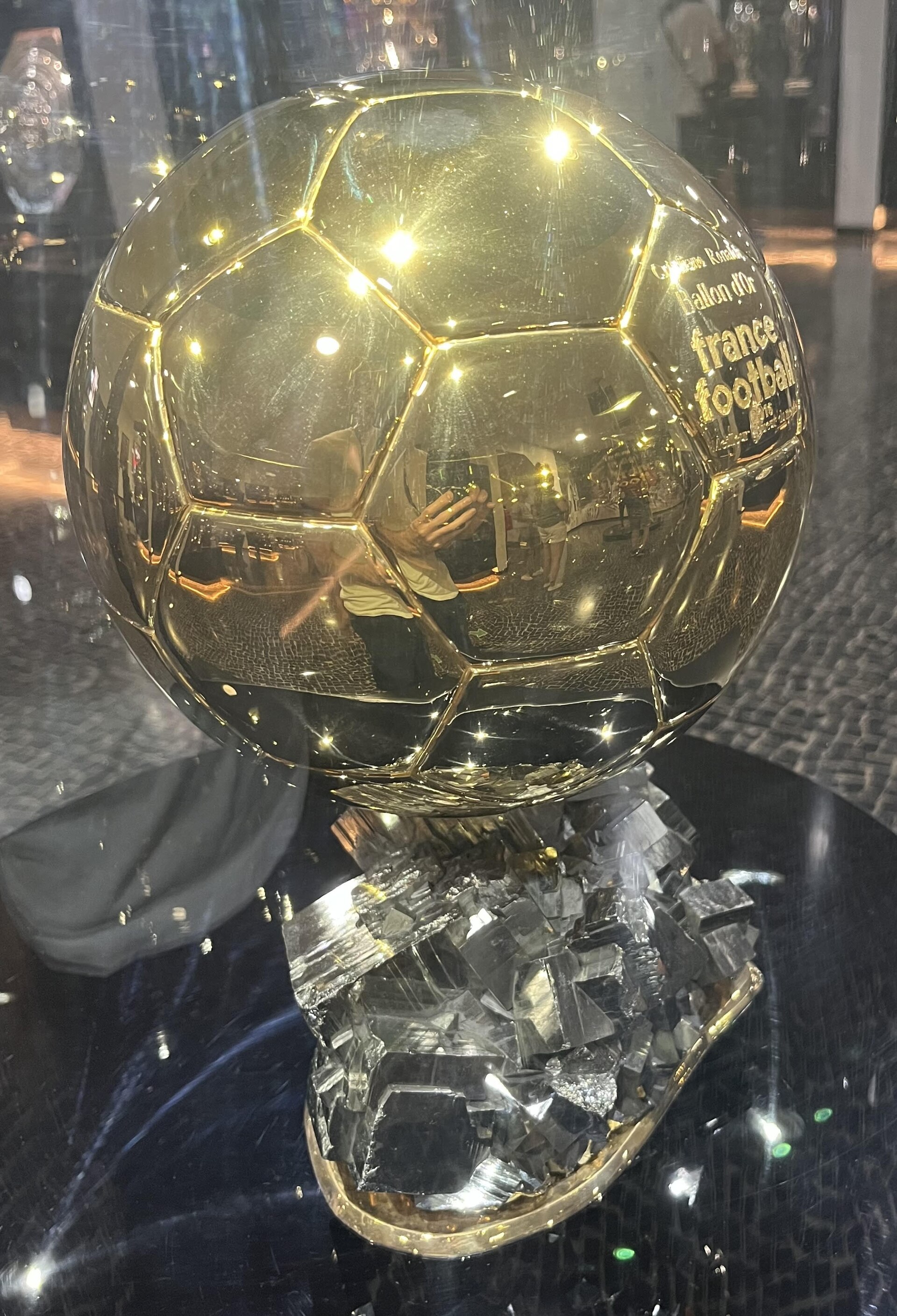 A close-up of the Ballon d'Or trophy, a golden soccer ball on a crystalline base, with reflections of people and lights on its surface.