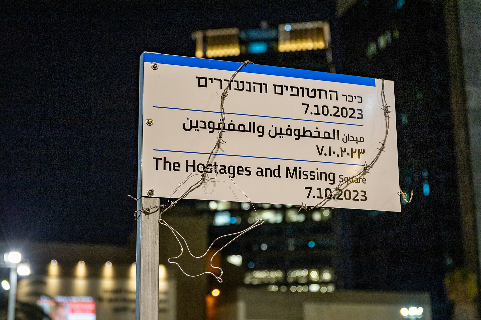 A sign reads "The Hostages and Missing Square" with dates in Hebrew and English, and barbed wire wrapped around it. A wire bird sculpture hangs from the barbed wire.