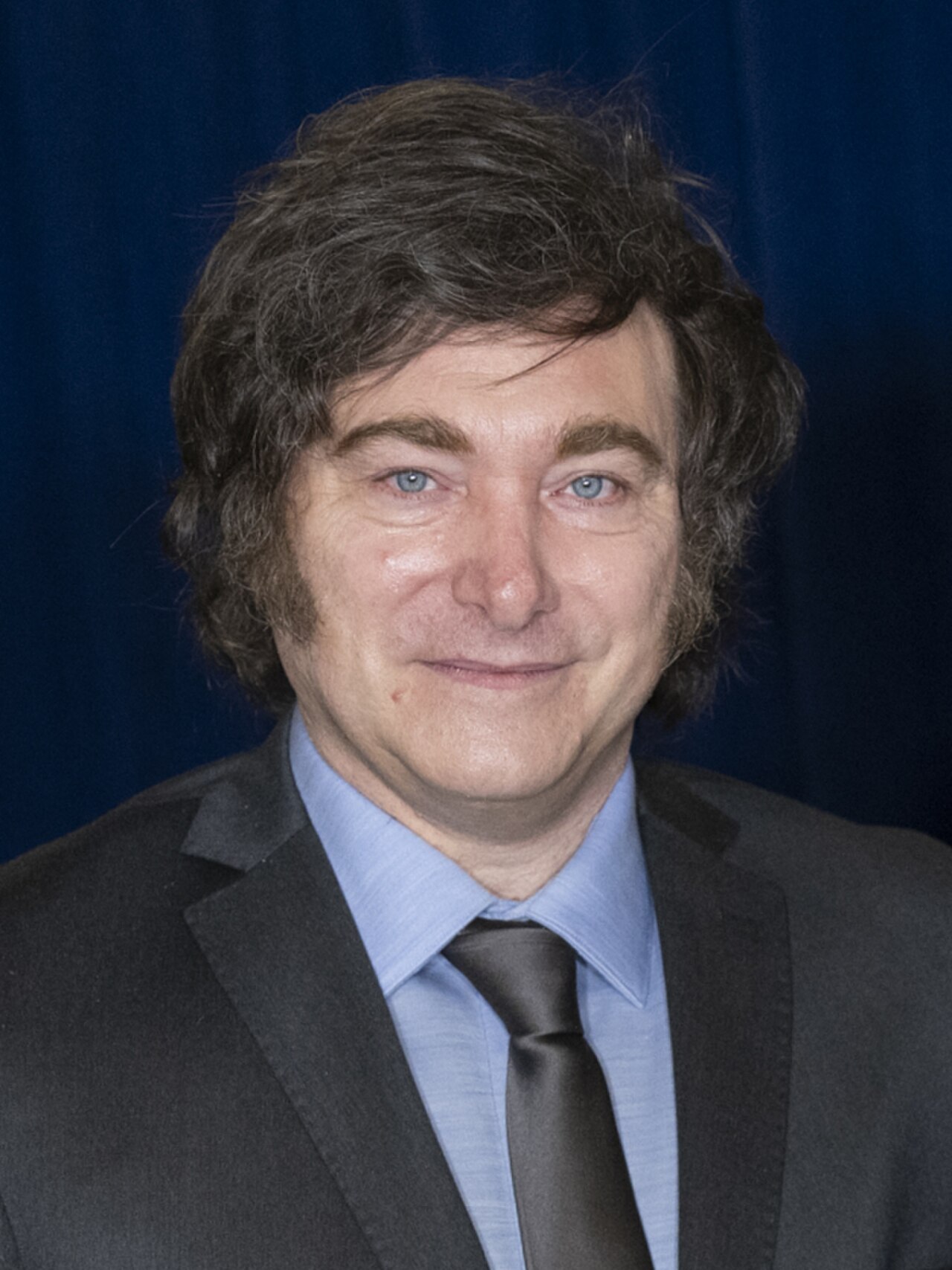 Head and shoulders of a middle-aged man with dark hair, blue eyes, wearing a gray suit, light blue shirt, and dark tie, against a dark blue background