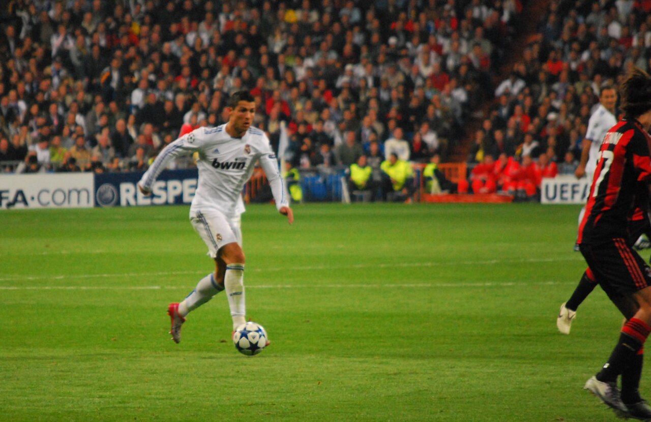 Cristiano Ronaldo in a white Real Madrid uniform dribbles a soccer ball on a green field in front of a crowd.