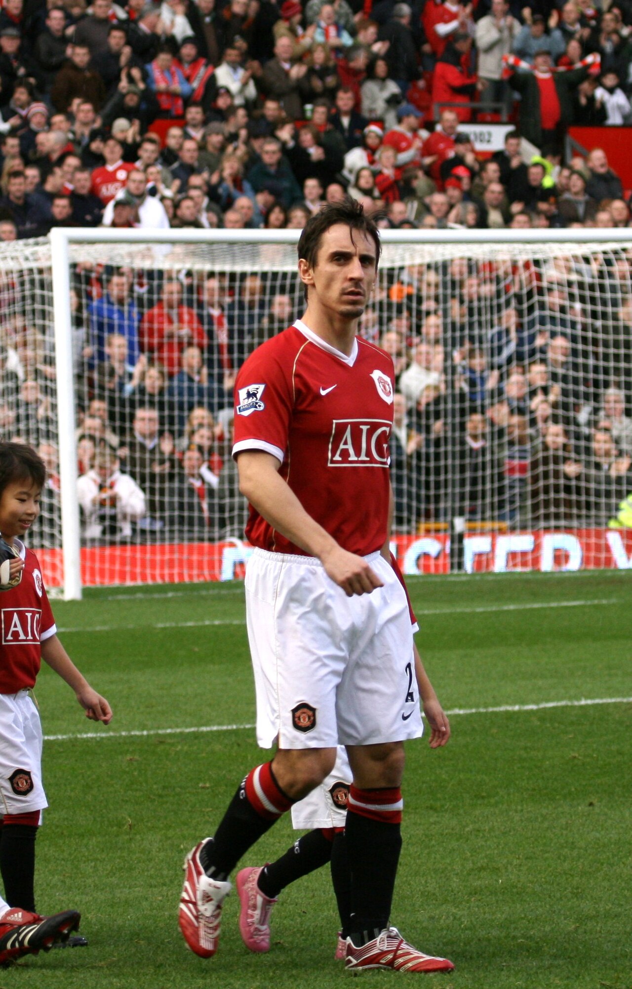 Gary Neville in a red Manchester United jersey and white shorts walks on a soccer field with a crowd in the background.