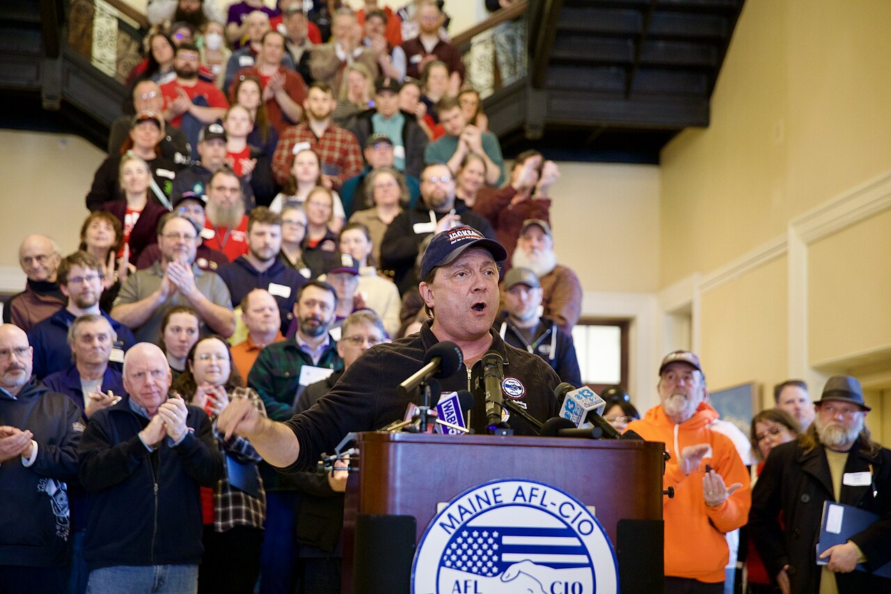 A man in a black shirt and baseball cap speaks at a podium with a Maine AFL-CIO logo, in front of a crowd of people.