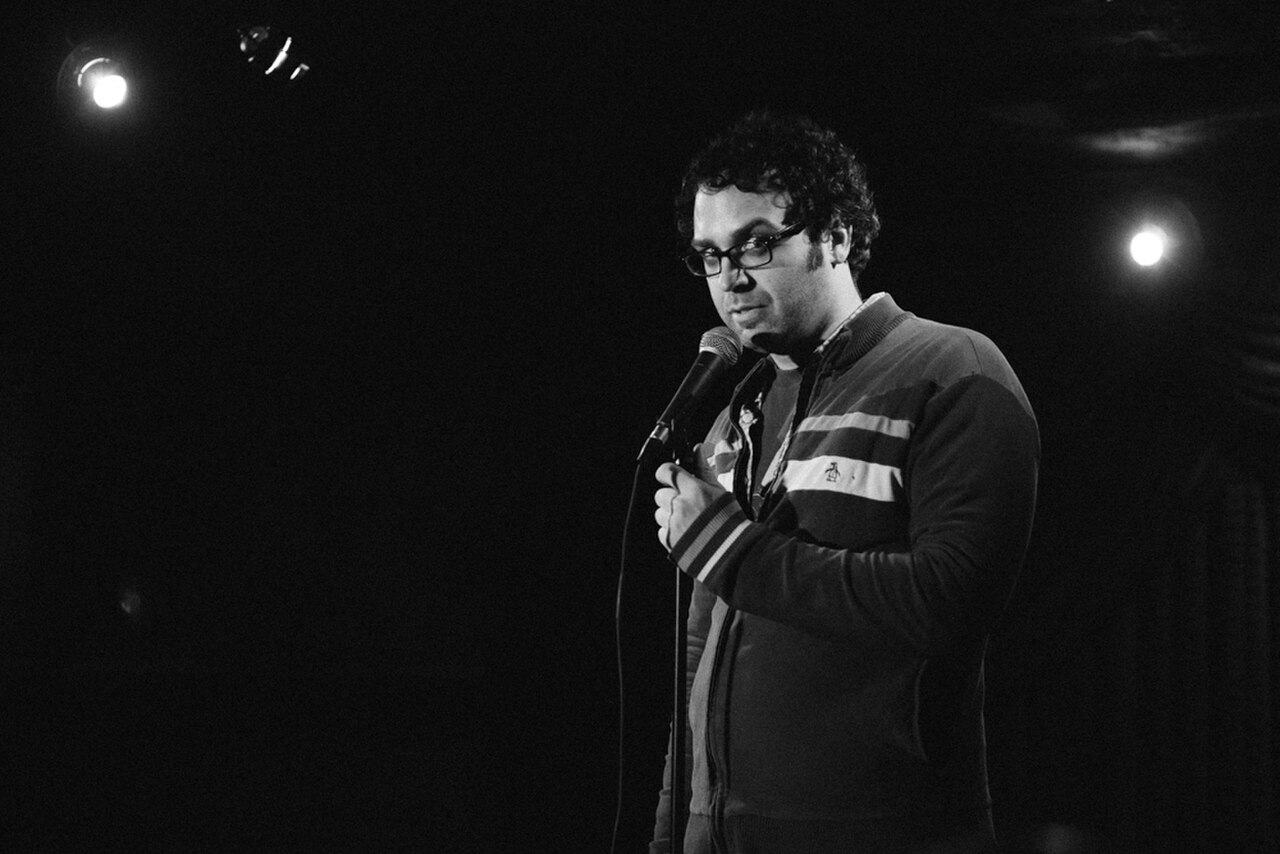 A man with curly hair and glasses stands on a stage holding a microphone, wearing a striped jacket.
