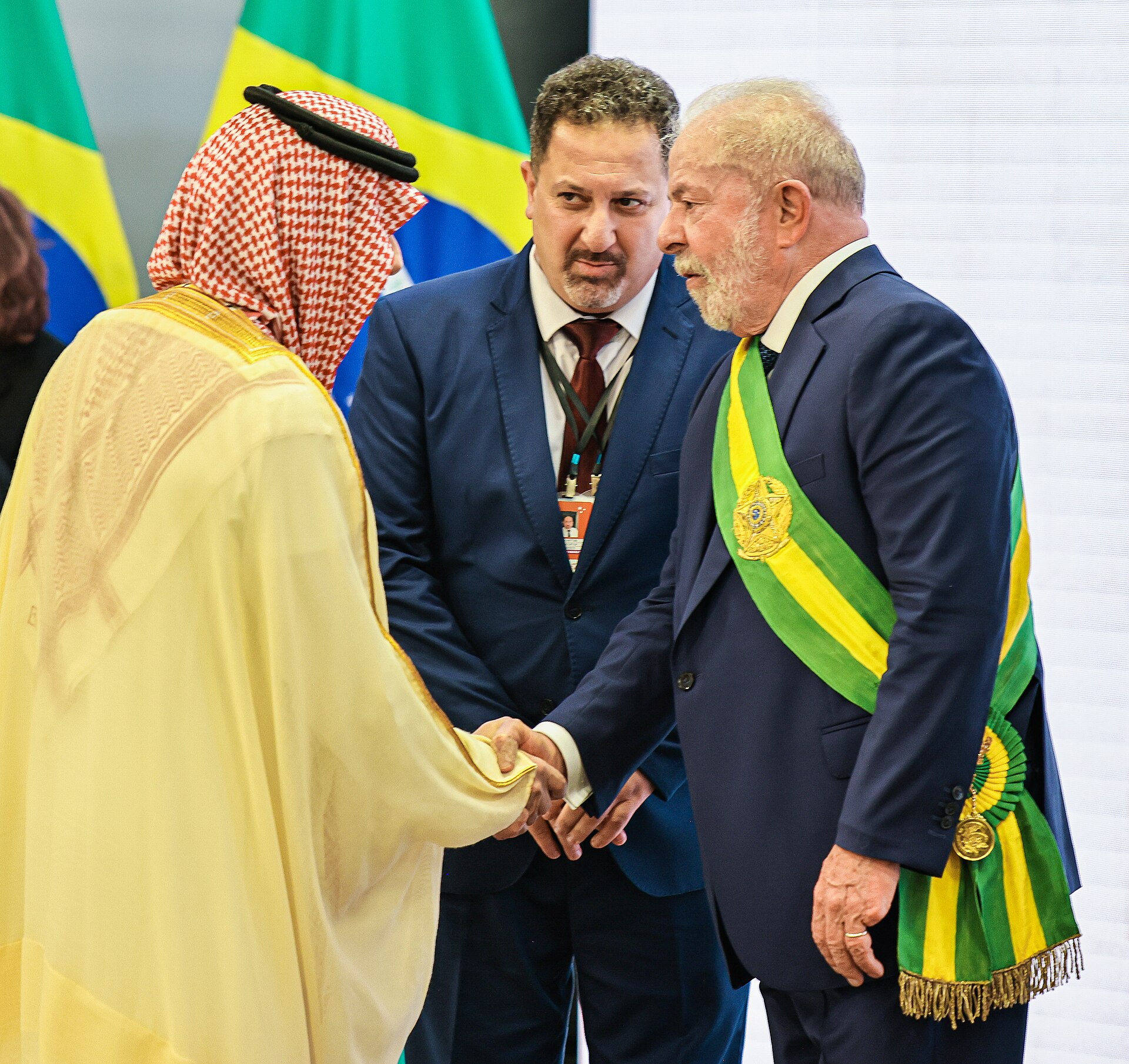 Luiz Inacio Lula da Silva in a blue suit and green and yellow sash shaking hands with a man in a white robe and headscarf