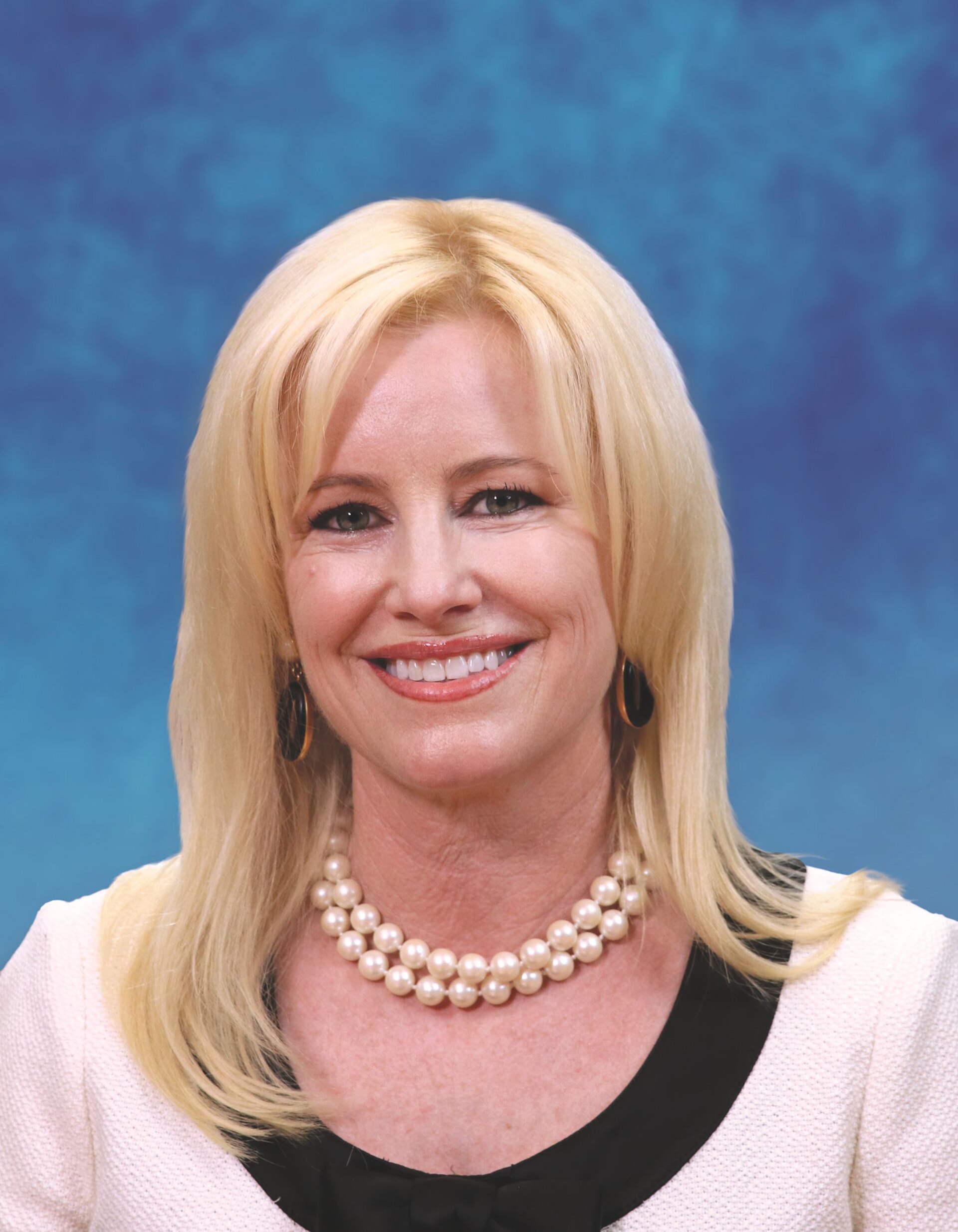 A blonde woman in a white jacket and pearl necklace smiles at the camera.