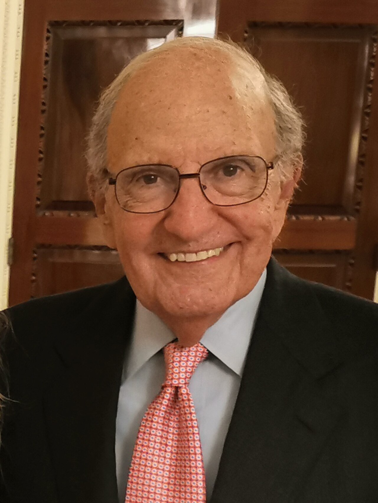 A smiling older man with glasses, wearing a black suit, light blue shirt, and red tie with a white pattern.