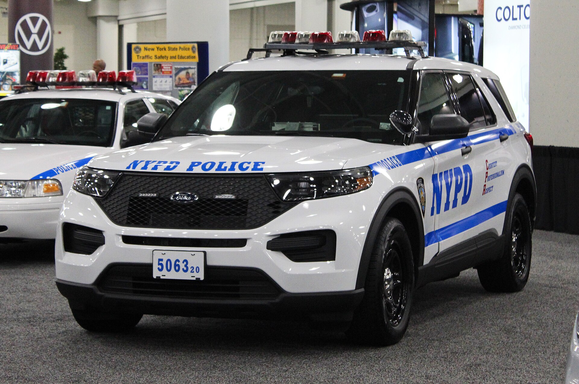 A white NYPD police SUV with blue lettering and stripes is parked indoors. The license plate reads 506320. Another white police car is partially visible to the left.