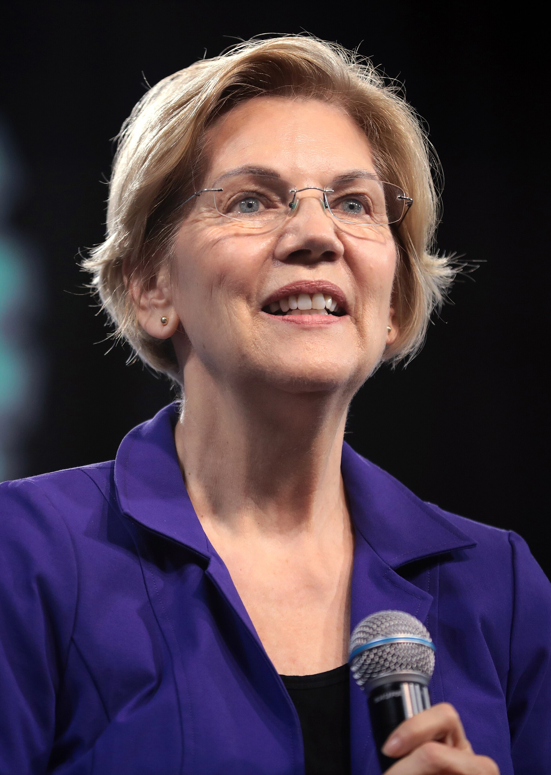 Elizabeth Warren from the shoulders up, wearing a purple blazer and holding a microphone