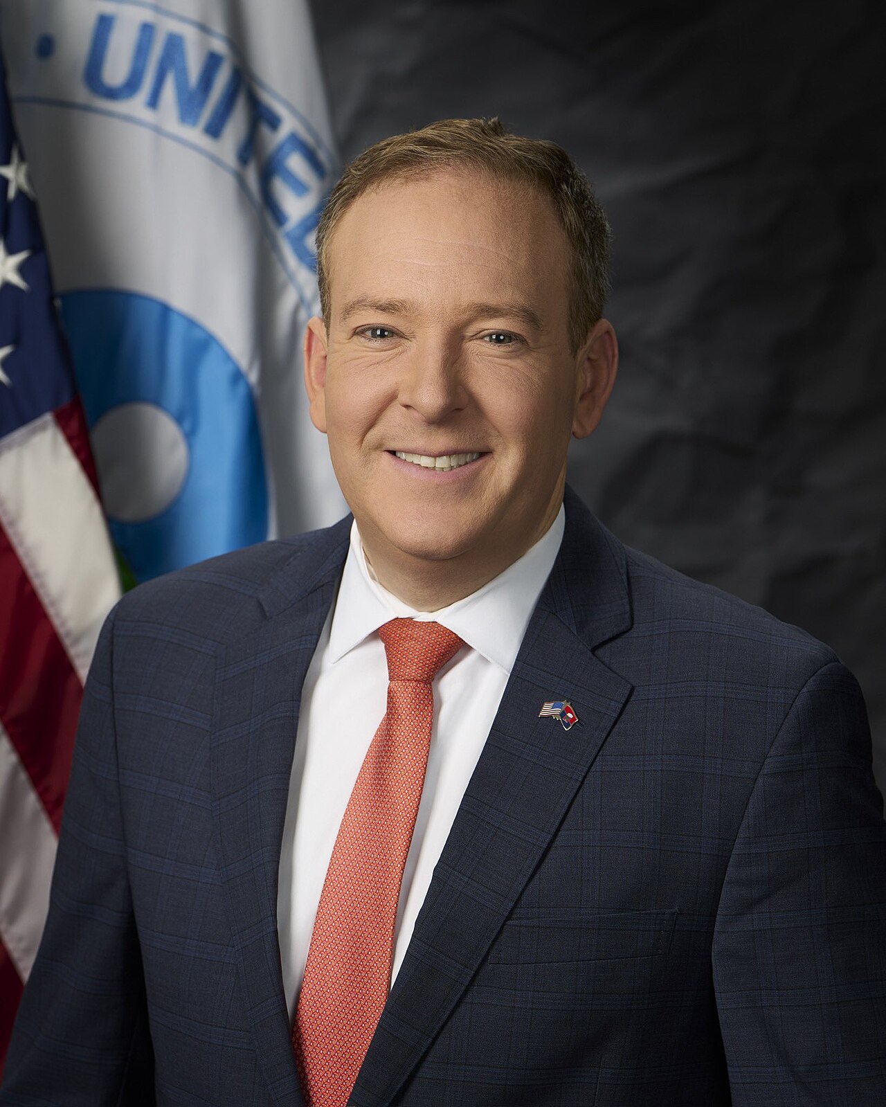 A portrait of Lee Zeldin, wearing a navy blue plaid suit, a white shirt, and an orange tie. He is smiling and looking at the camera. Behind him, to the left, is the American flag and a flag with a blue circle and the word "UNITED" visible.