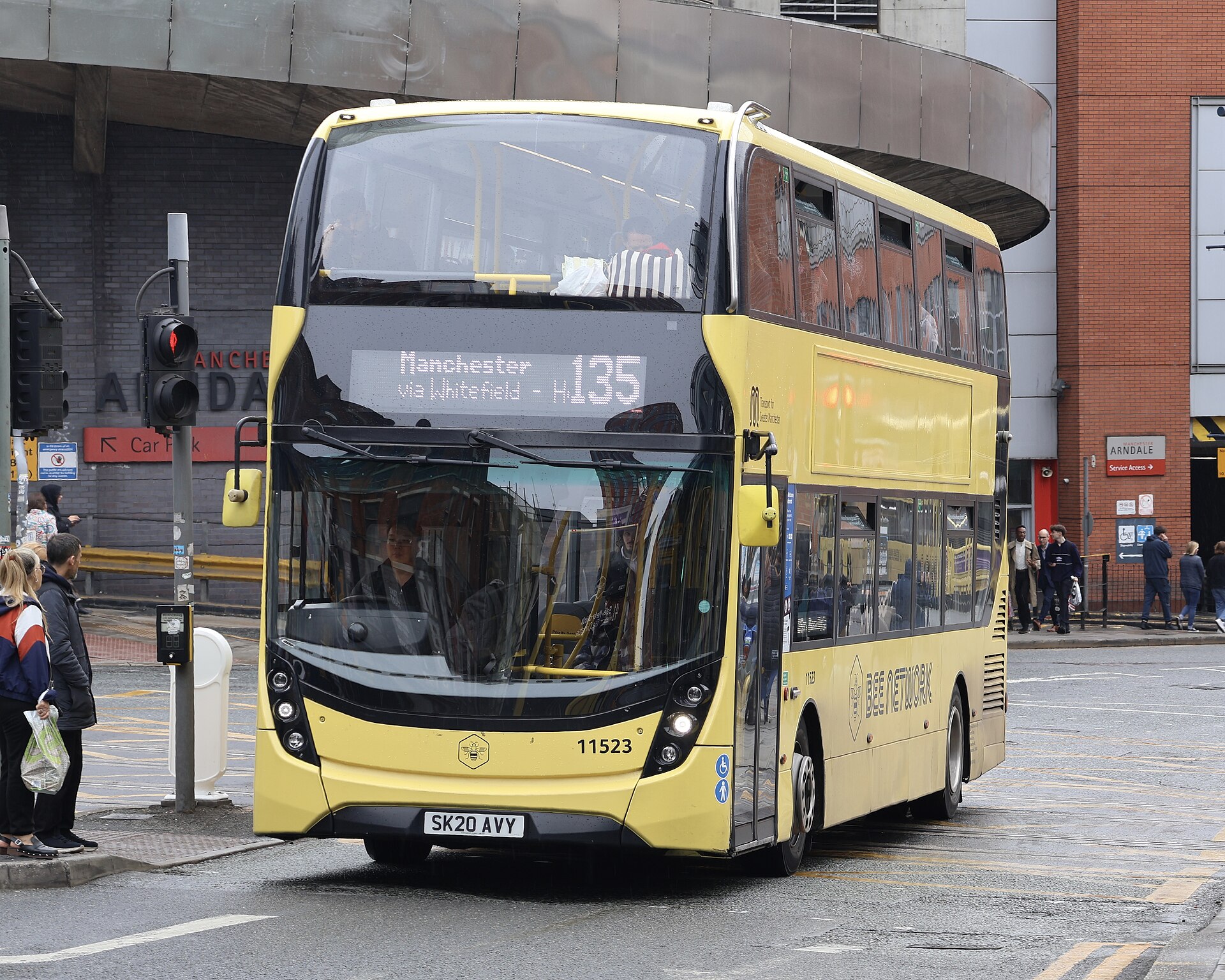 A yellow double-decker bus with "Bee Network" written on its side is driving on a wet street. The bus displays "Manchester via Whitefield - H135" on its front. People are visible inside the bus and on the sidewalk.