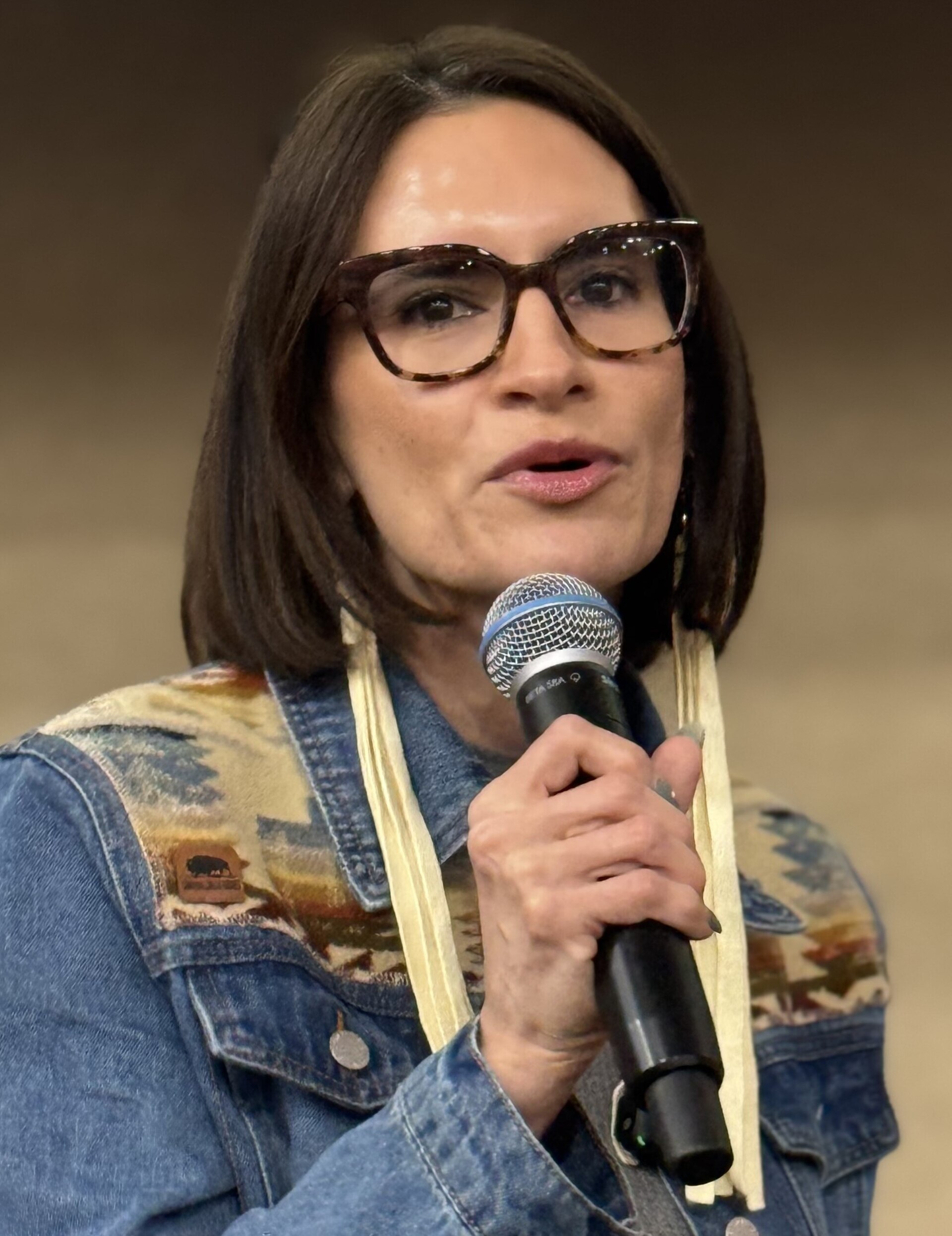 A woman with dark hair and glasses speaks into a microphone, wearing a denim jacket with a patterned yoke and light-colored fringe.