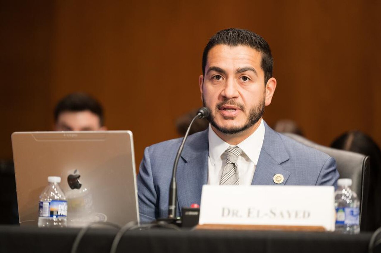 A man with short dark hair and a beard wearing a blue suit and tie sits at a table with a microphone, laptop, and name card that reads Dr. El-Sayed.