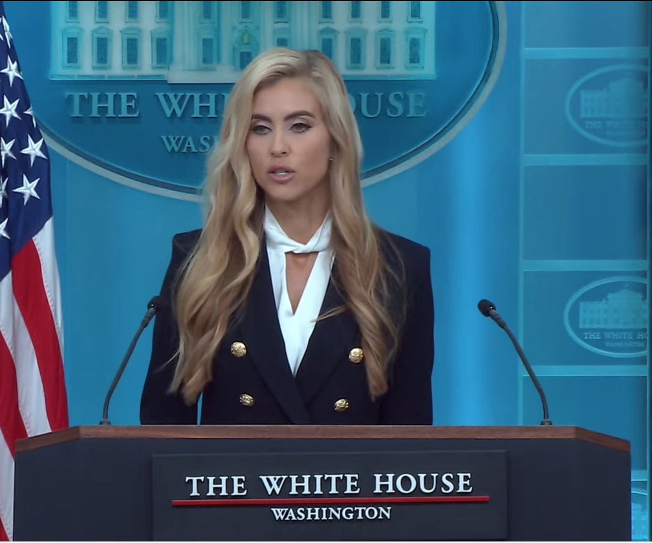 A blonde woman in a black blazer with gold buttons and a white blouse stands at a podium with two microphones, in front of a blue backdrop with the White House logo and an American flag to her left.