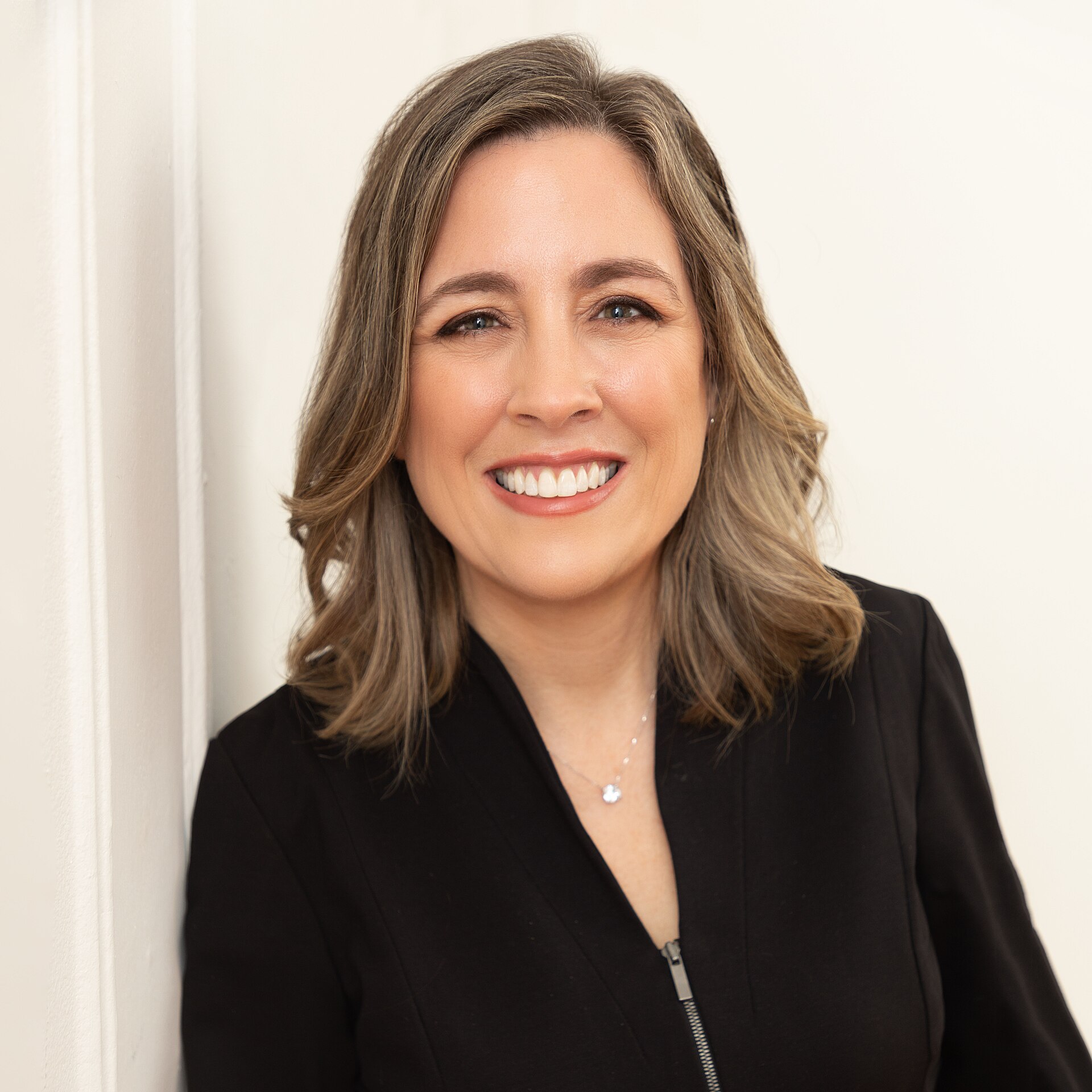 A smiling middle-aged woman with shoulder-length brown hair and a black jacket, leaning against a white wall.