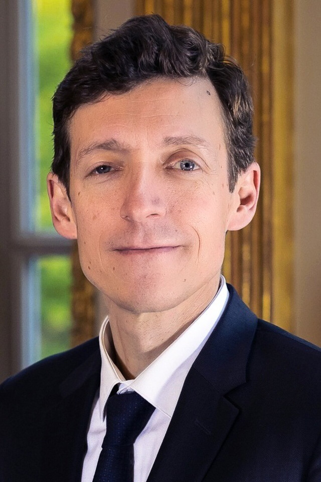 A man with short dark hair, wearing a dark suit, white shirt, and dark tie, looking at the camera.