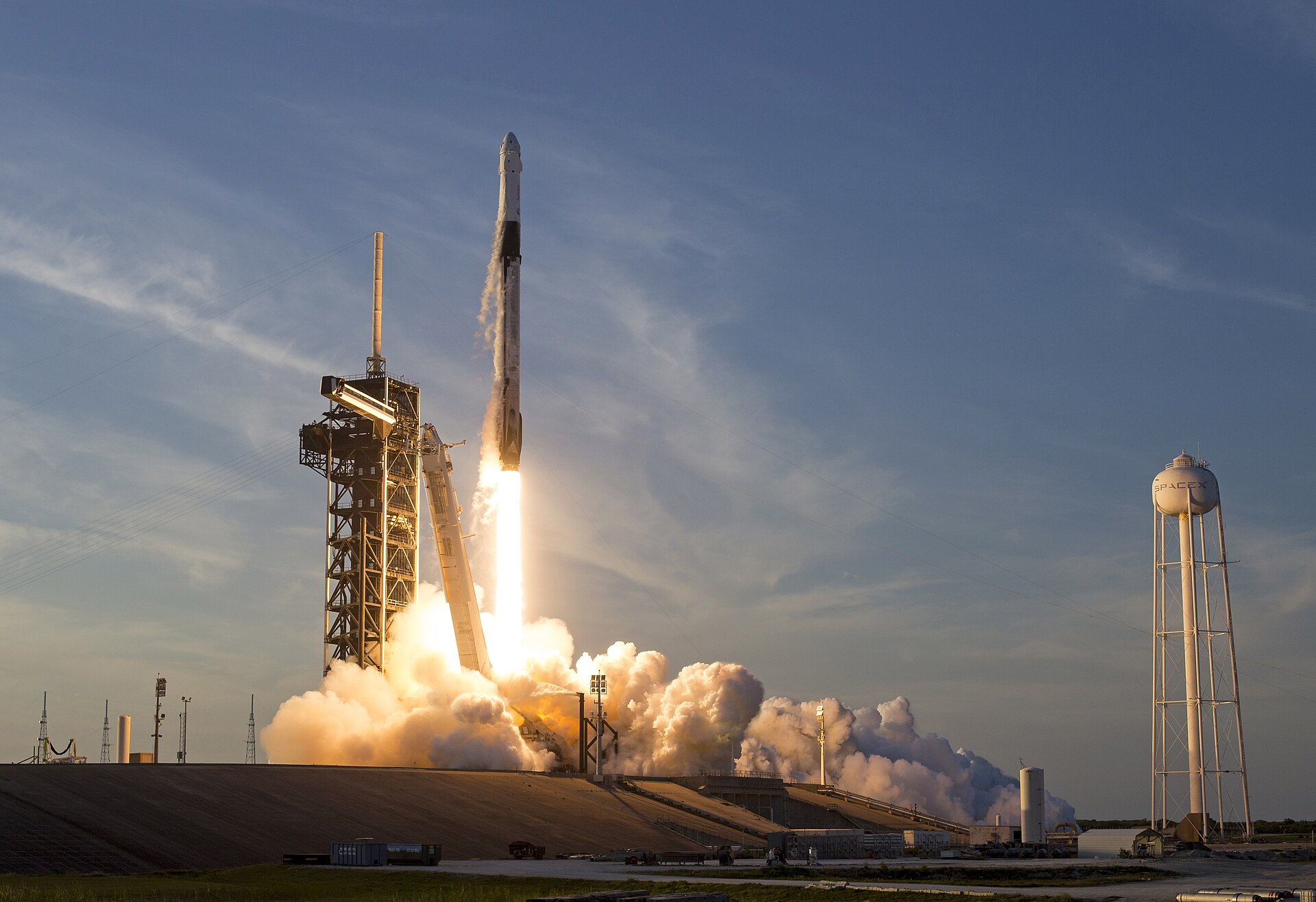 A SpaceX rocket launches from a launchpad, with a SpaceX water tower visible in the background.