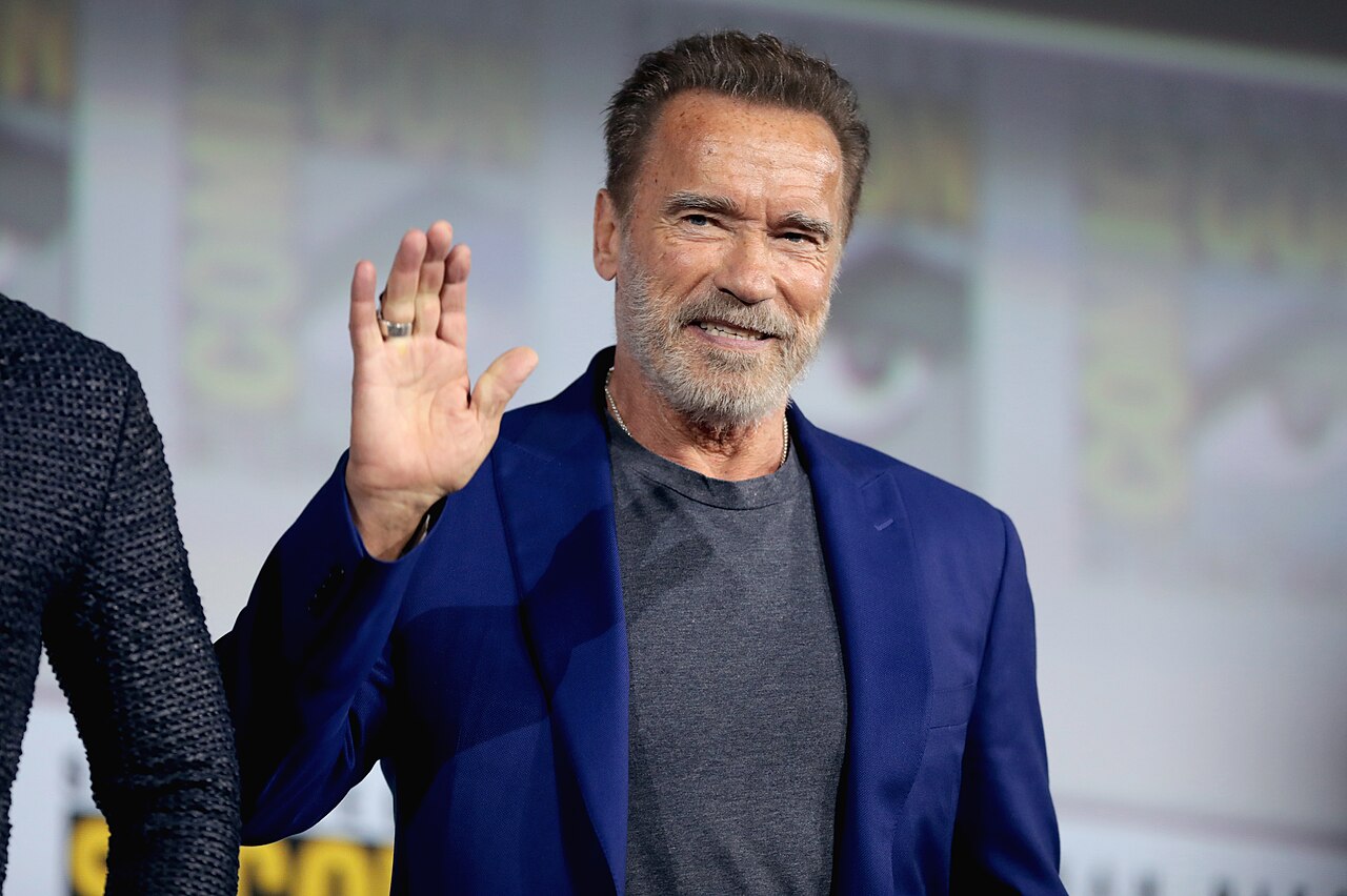 Arnold Schwarzenegger waving on stage at San Diego Comic-Con in 2019, wearing a blue jacket.