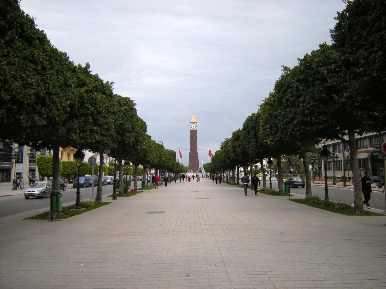 A wide pedestrian walkway lined with trees leads towards a tall clock tower in the distance. Cars are visible on the street to the left, and buildings line the right side of the street.