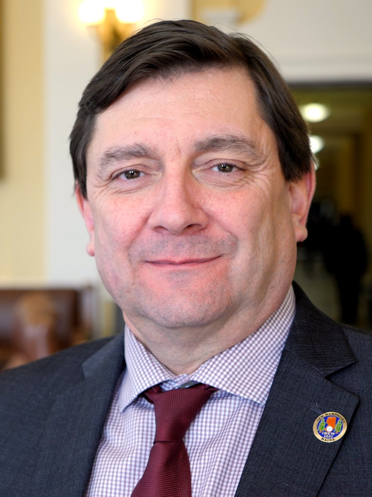 A man wearing a suit, a checkered shirt, and a maroon tie smiles at the camera. He has a pin on his lapel that says "New Hampshire 1630 Maine."
