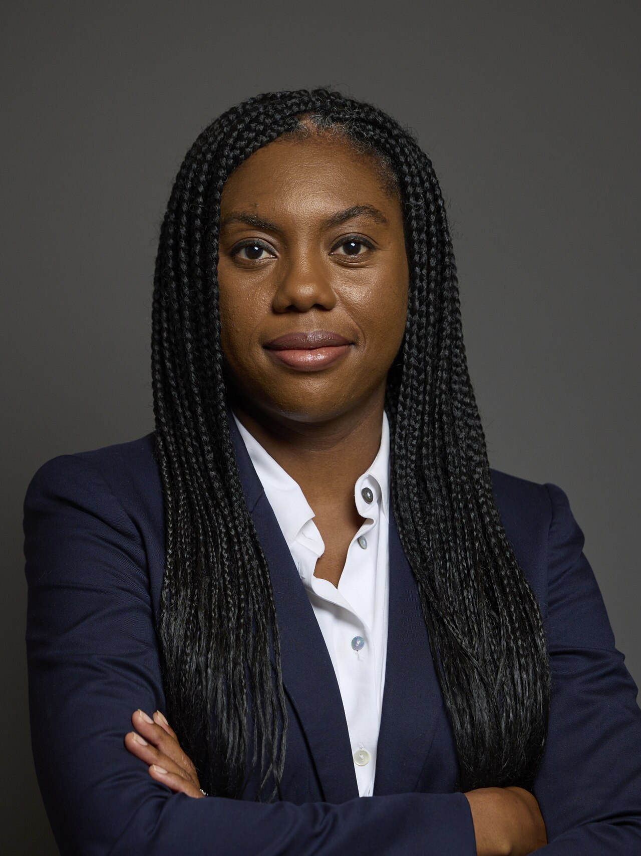 Kemi Badenoch, a black woman with long braids, wearing a white shirt and blue blazer, looking at the camera with her arms crossed.