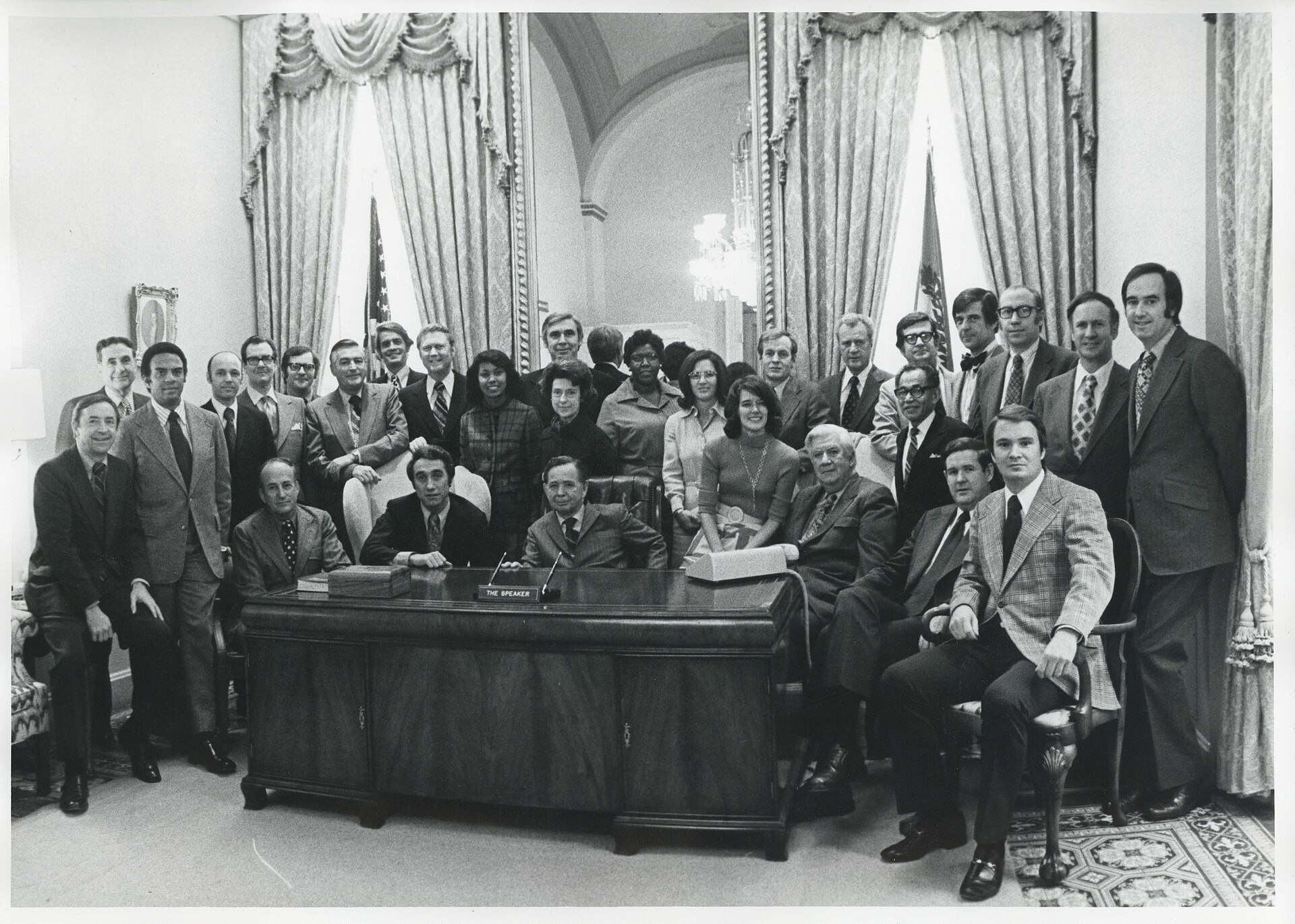 A group of people, mostly men in suits, pose for a photograph in a formal room with ornate curtains and a large mirror.